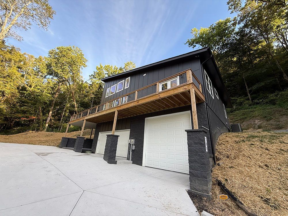 Black two-story house with white garage doors and a deck. Driveway slopes up to the house.
