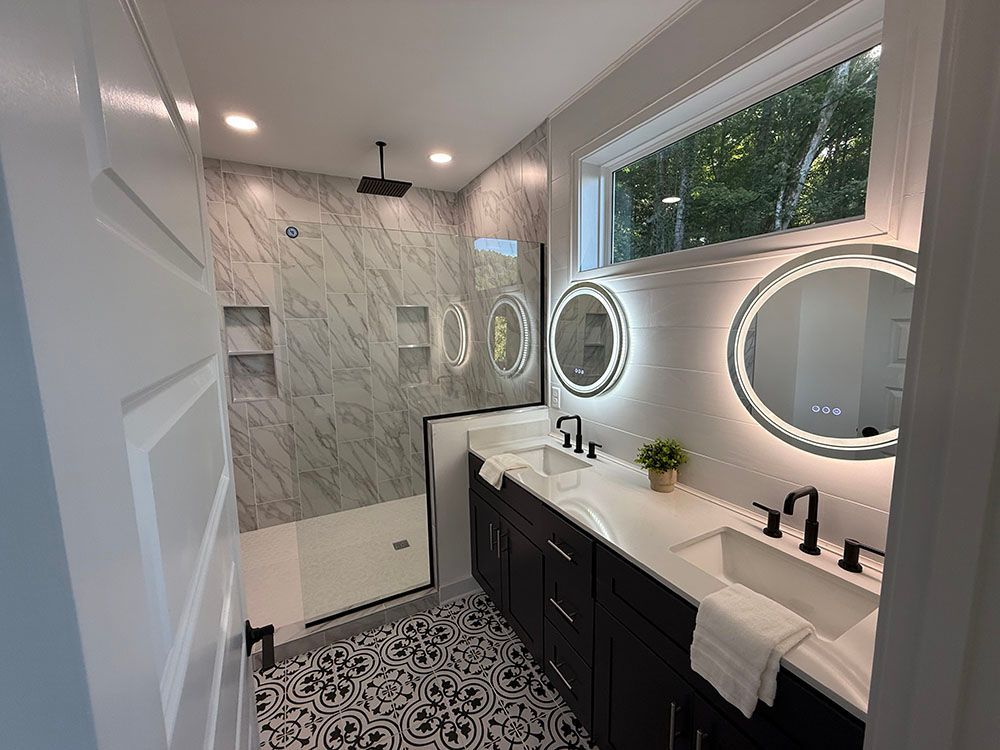 Modern bathroom with marble shower, black vanity, patterned floor, and round mirrors with lights.