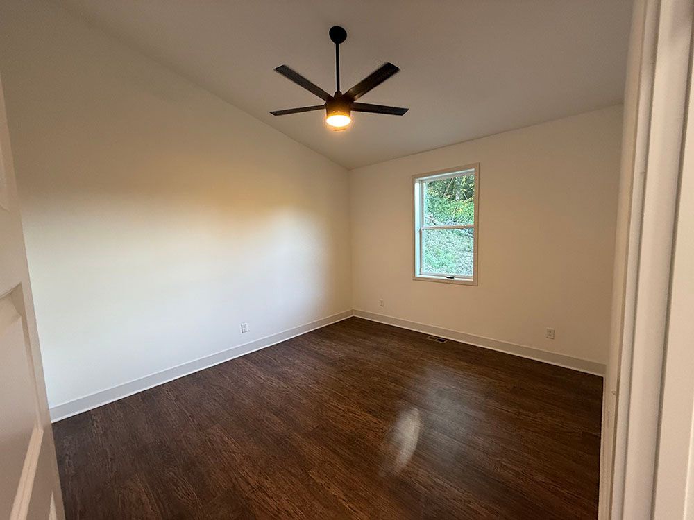 Empty bedroom with dark wood floor, white walls, a ceiling fan, and a window.
