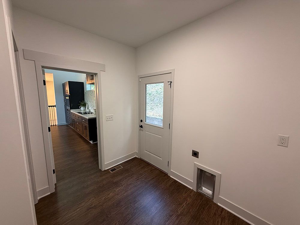 A white-walled hallway with wood floors, a door, and a doorway leading to a kitchen.