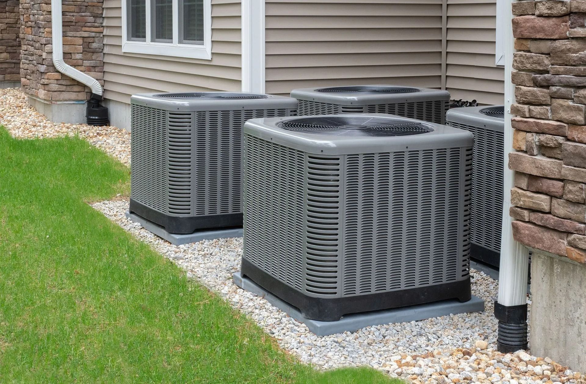 Four air conditioning units on gravel next to a house with beige siding and a stone wall.