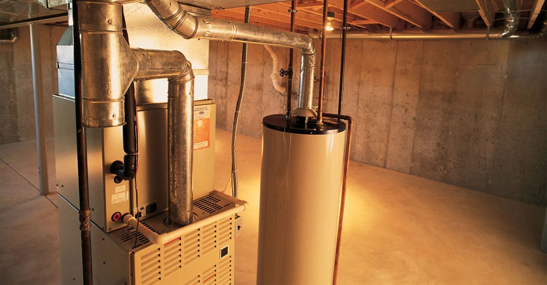 Furnace and water heater in a basement. Beige appliances with silver ductwork. Concrete walls and floor.