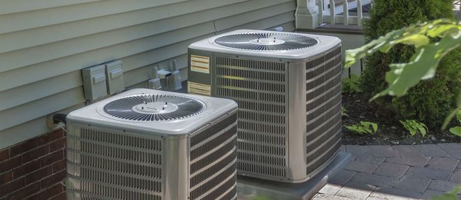 Two air conditioning units sit on a patio next to a house with brick and siding.