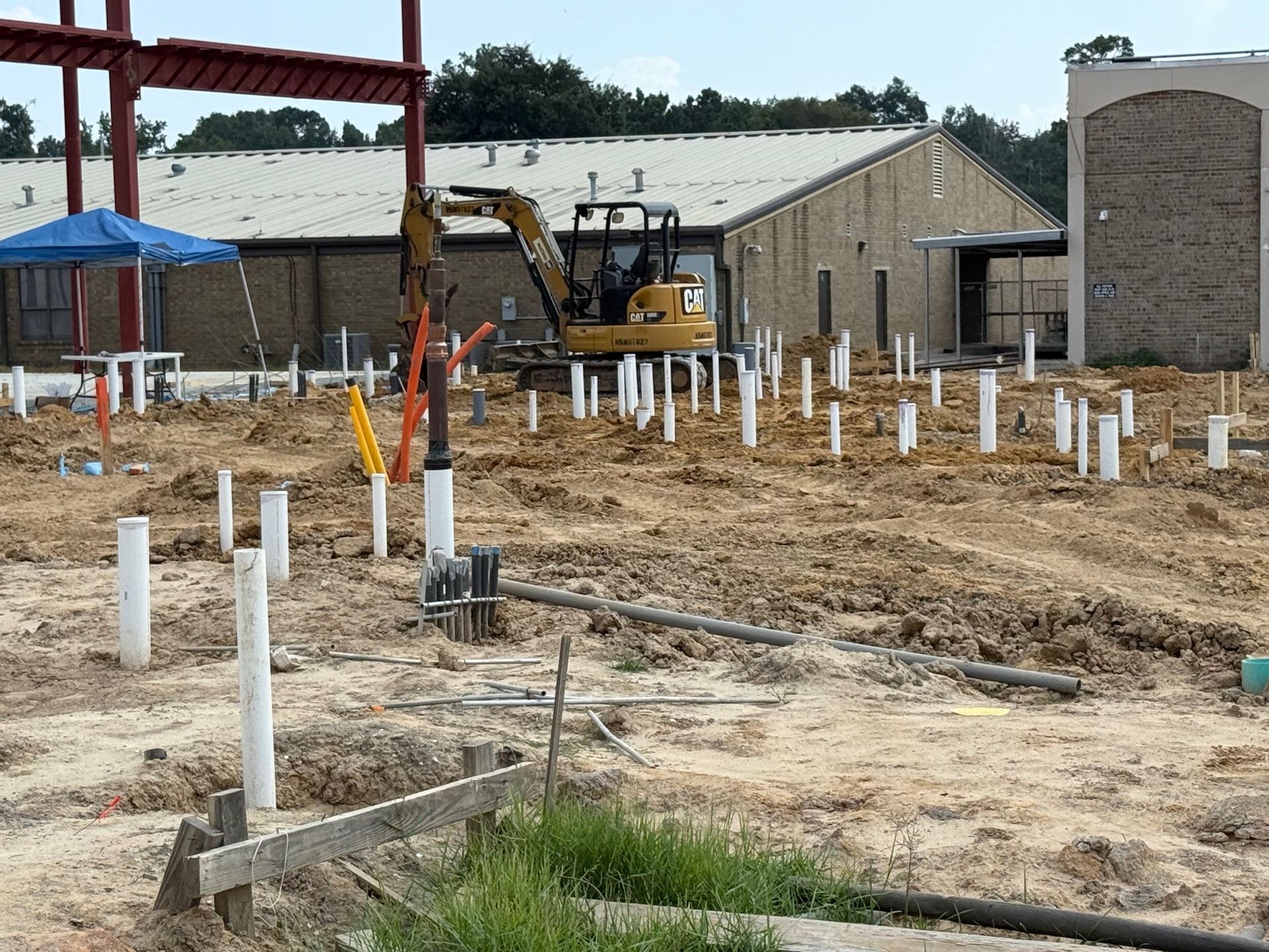 Construction site with pipes in the ground, excavator, and building in the background.