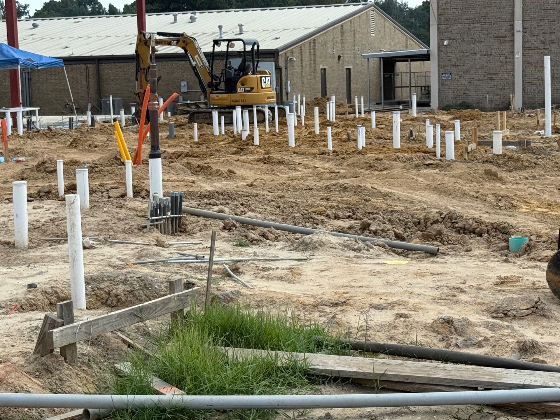 Construction site with a backhoe, white pipes, and dirt near a building.