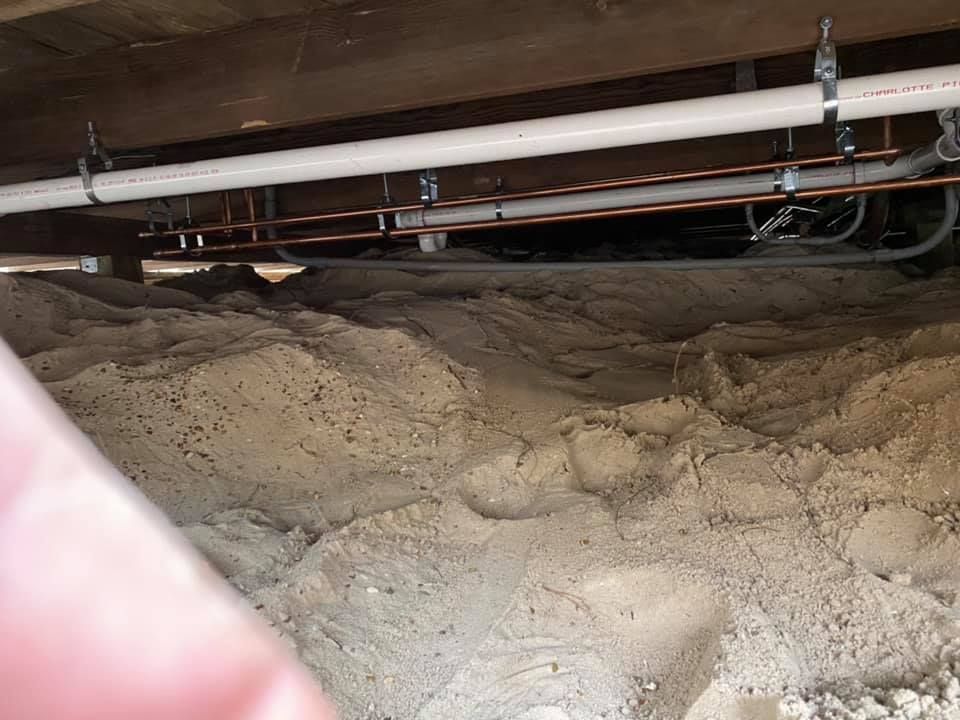 Under-house view: Sand-covered ground, with pipes and wiring attached to the wooden floor above.