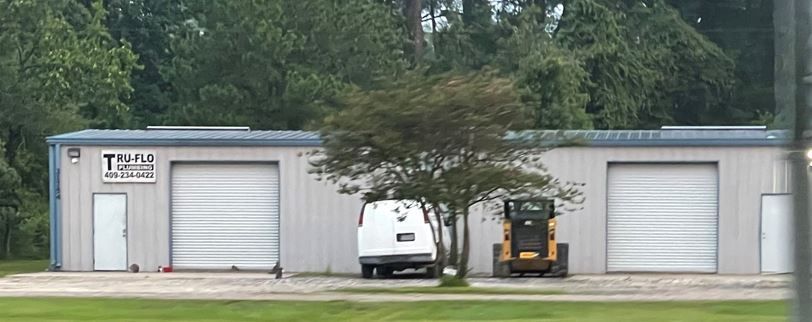 A commercial building with two garage doors, a white van, a skid steer, and a tree in front.