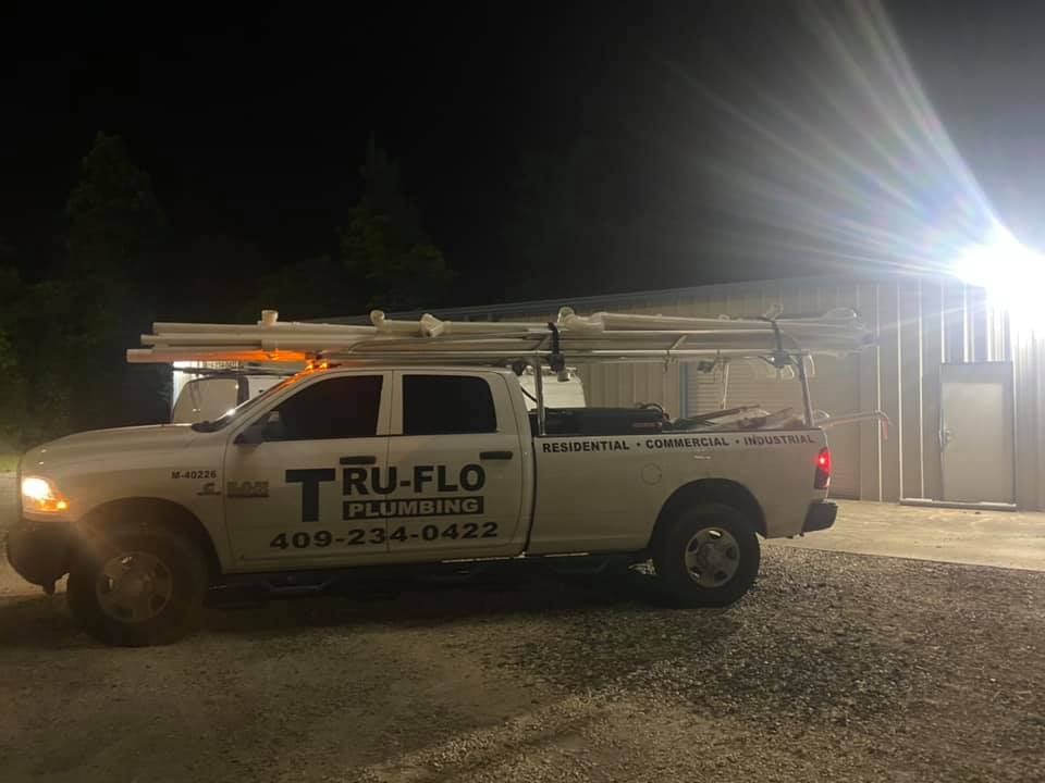 White Tru-Flo Plumbing truck parked at night, carrying pipes on top, near a building.