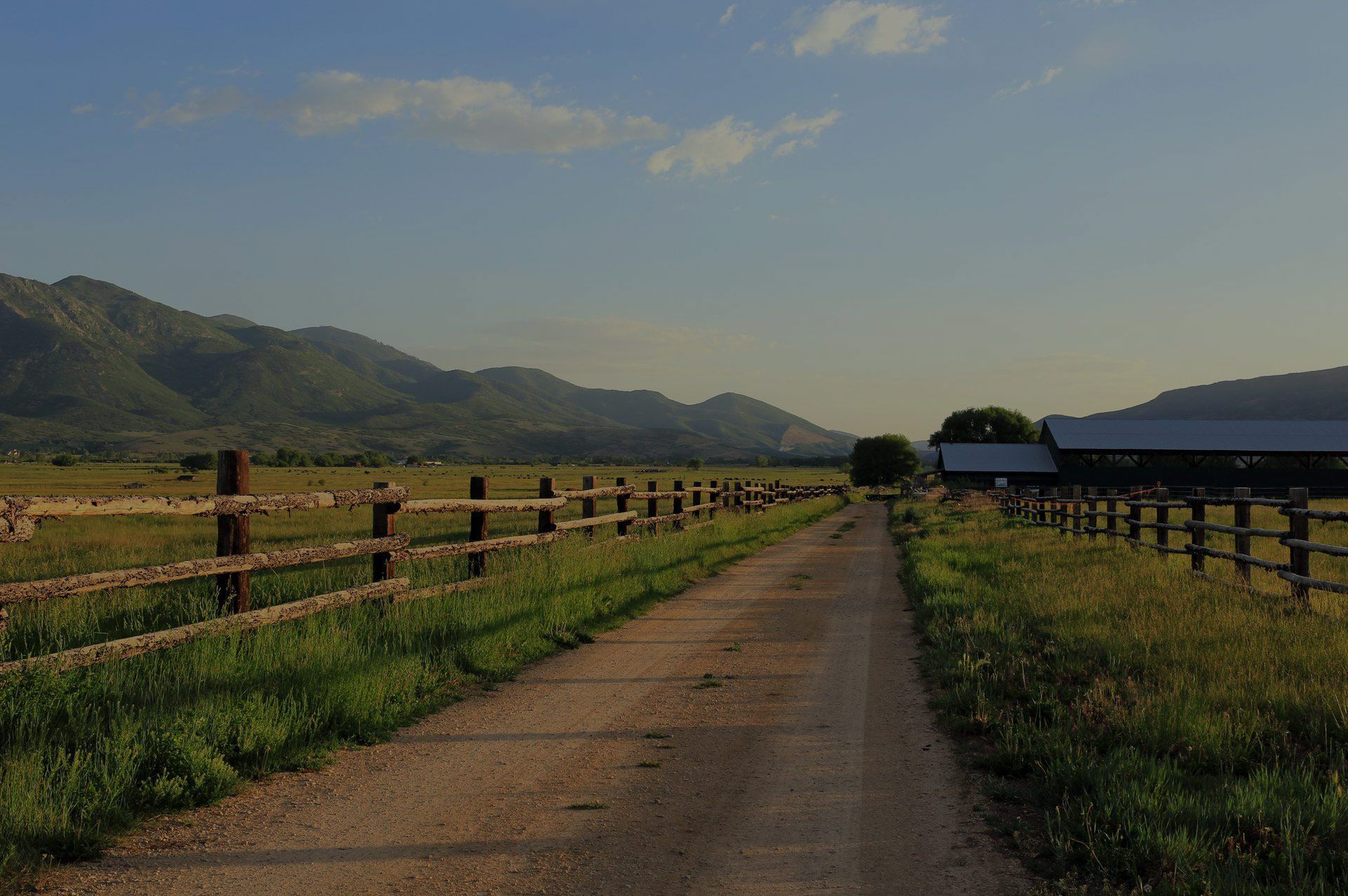 Dirt road through a grassy field, wooden fences on both sides, leading towards a barn and mountains under a blue sky.