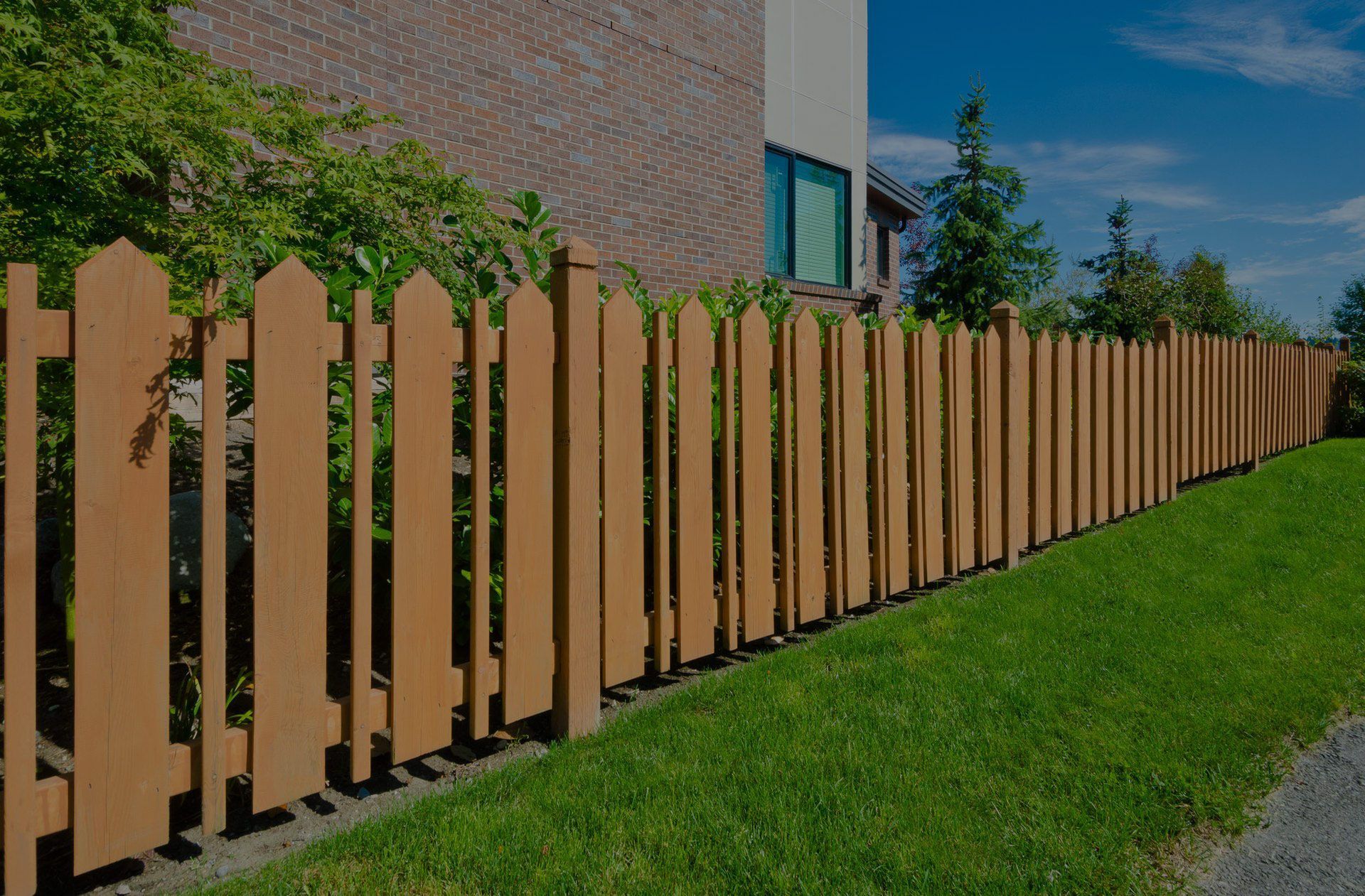 Wooden picket fence bordering a green lawn, with a brick building and trees in the background.