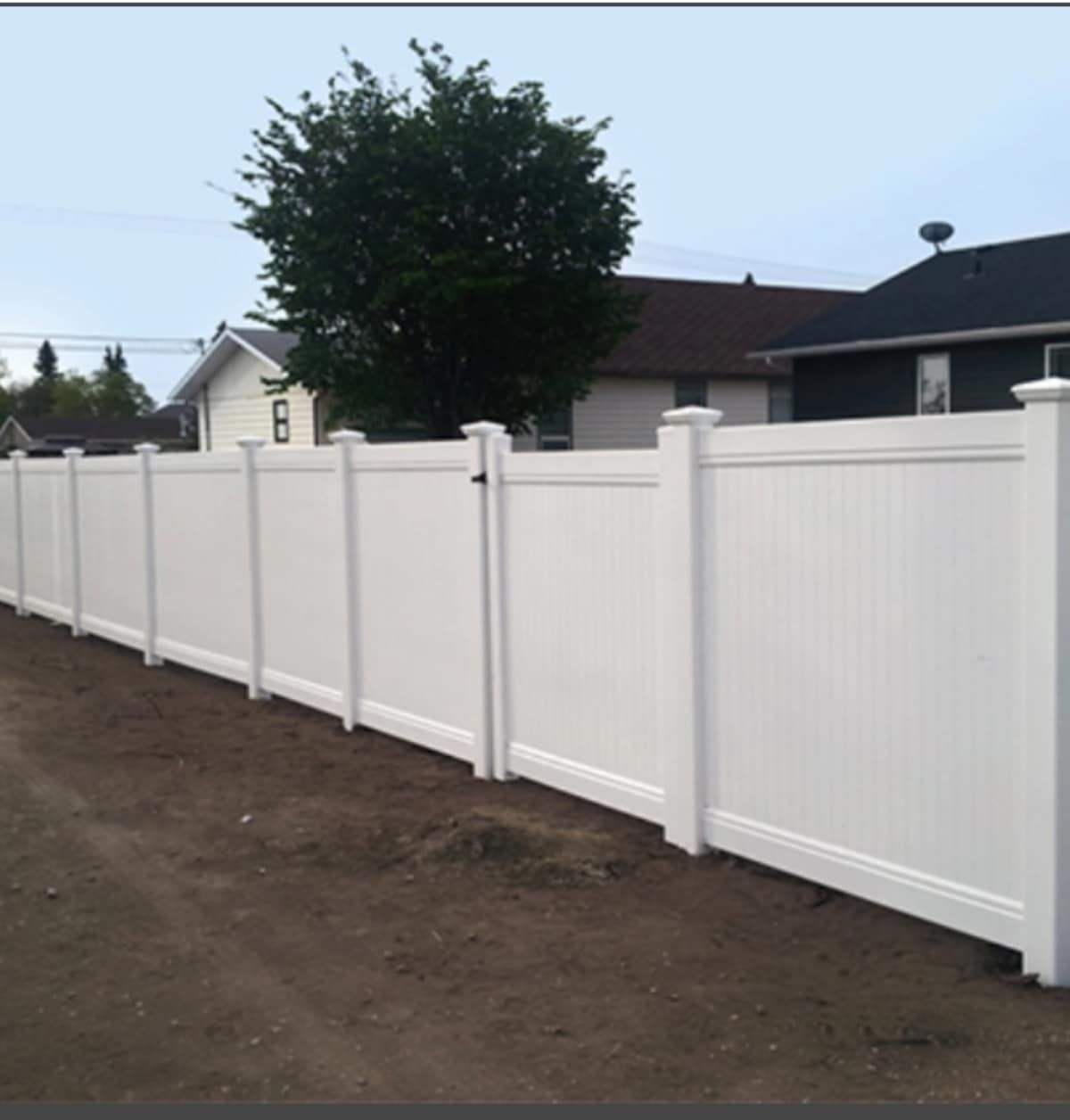 White vinyl fence enclosing a yard, with a gate, against a backdrop of houses and a tree under a cloudy sky.
