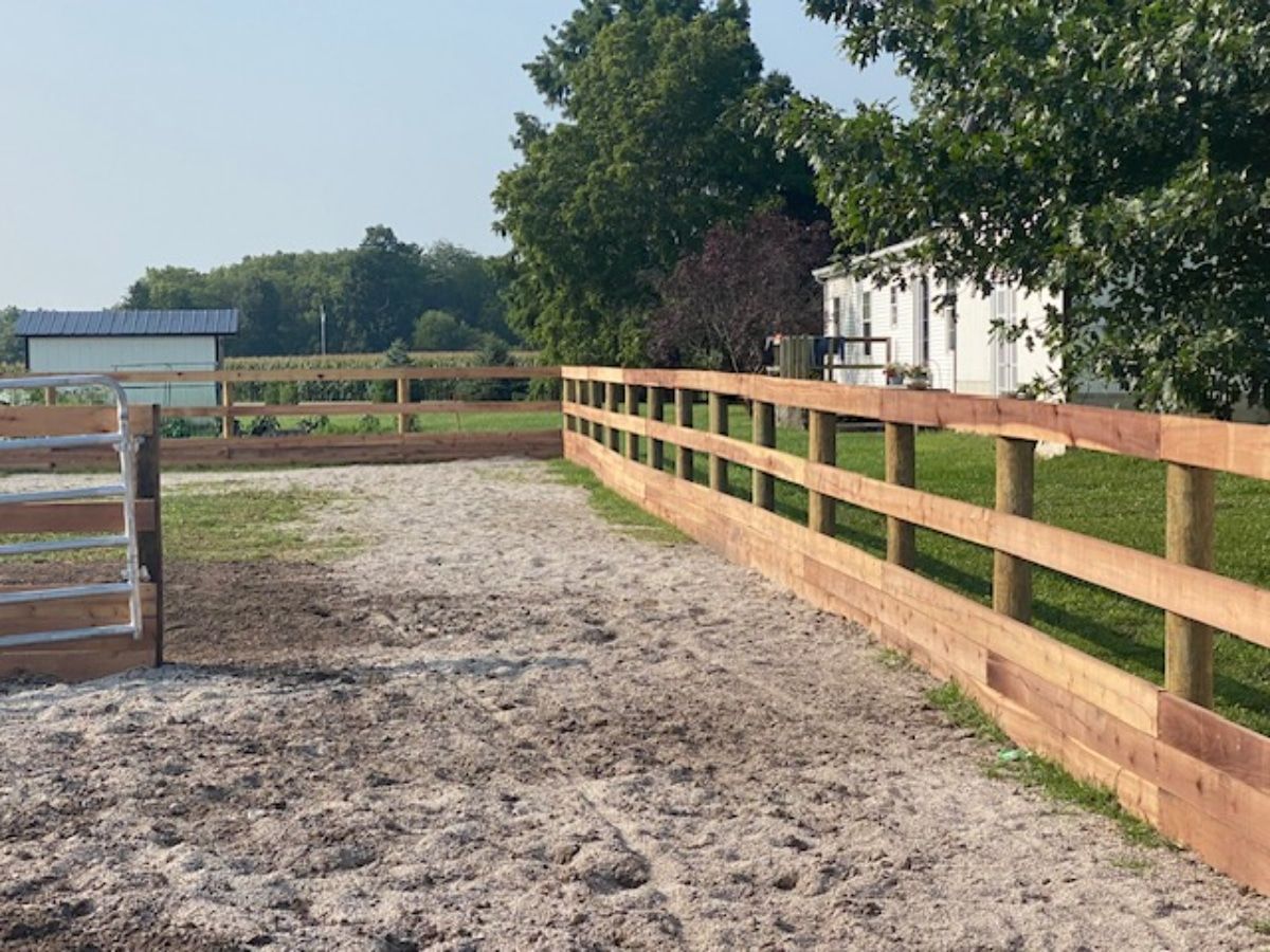 An empty horse riding arena enclosed by a wooden fence, with trees and a building in the background on a sunny day.