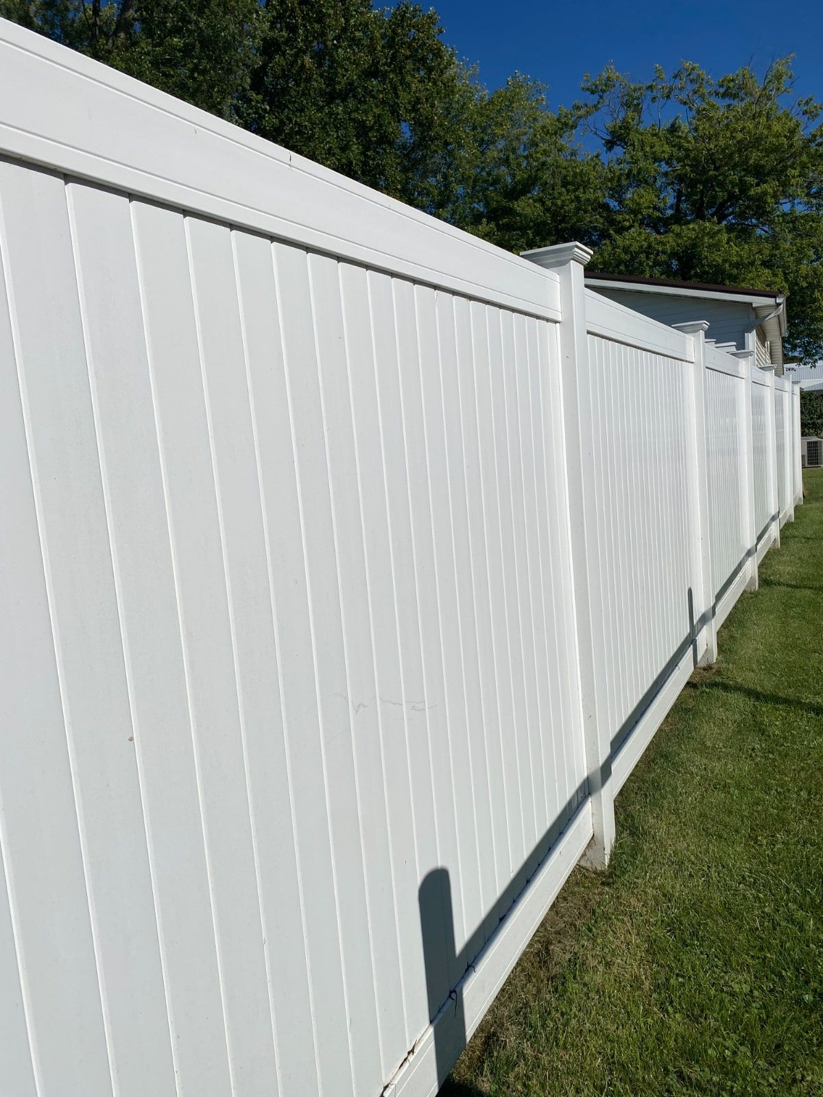 White vinyl fence extending along green grass on a sunny day. Trees are visible in the background.