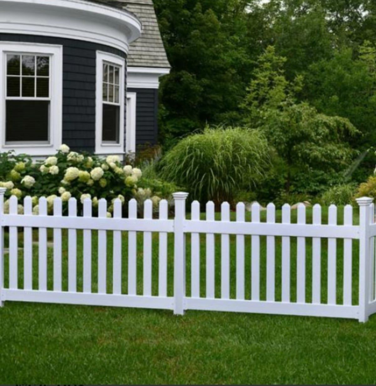 White picket fence in front of a dark house with green landscaping.