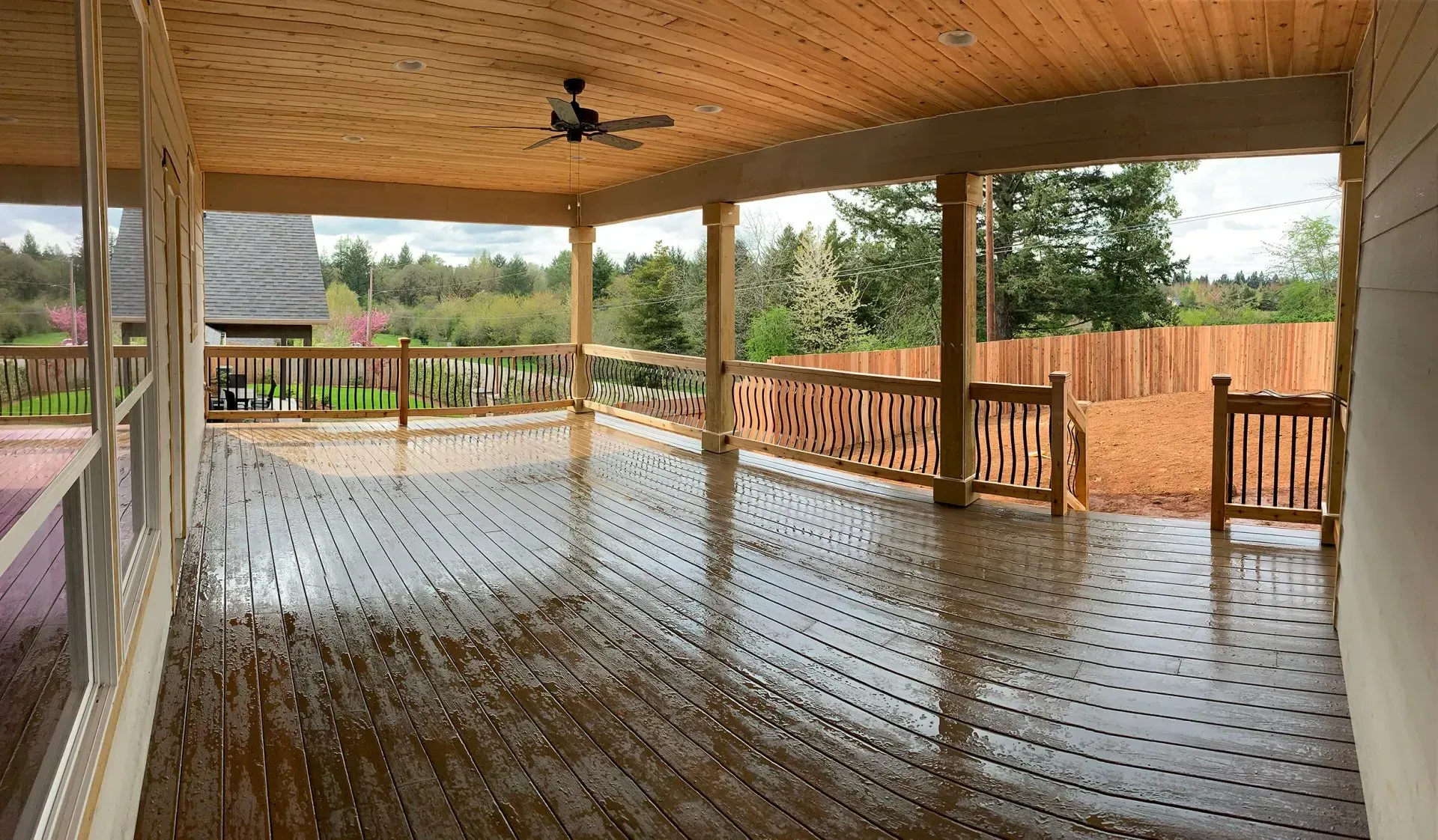 Covered wooden deck with a ceiling fan, overlooking a fenced yard and trees.