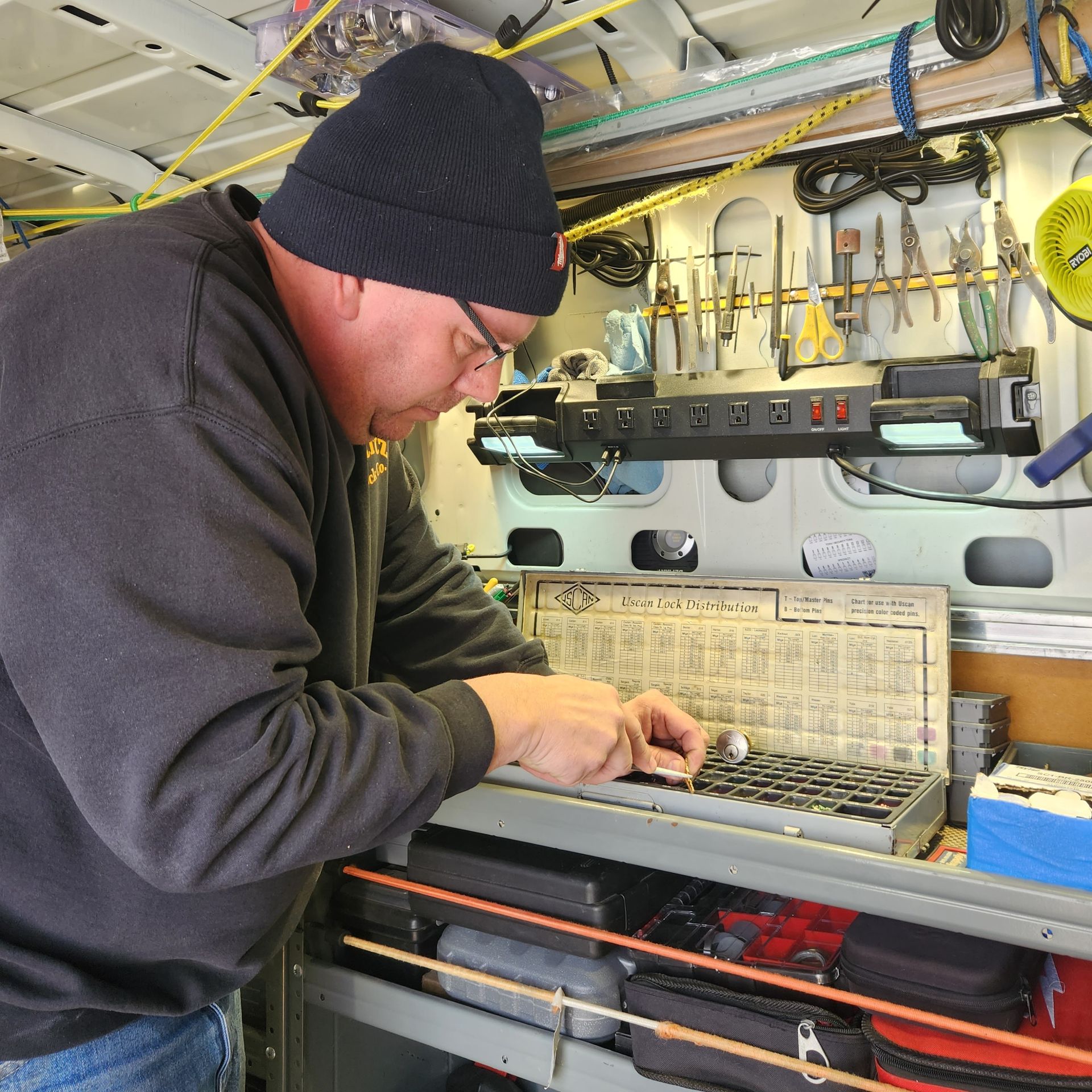 Man in a black beanie and glasses working on small parts, likely in a van with tools organized on the wall.