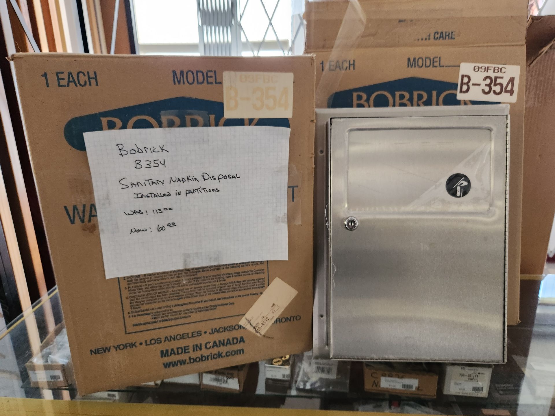 Stainless steel restroom sanitary napkin dispenser box with handwritten label.