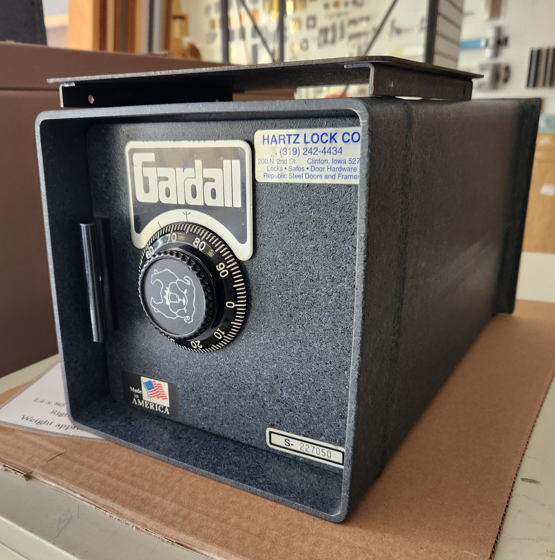 A gray Gardall safe with a combination dial, labeled with a Hartz Lock Co. sticker, sitting on a cardboard surface.