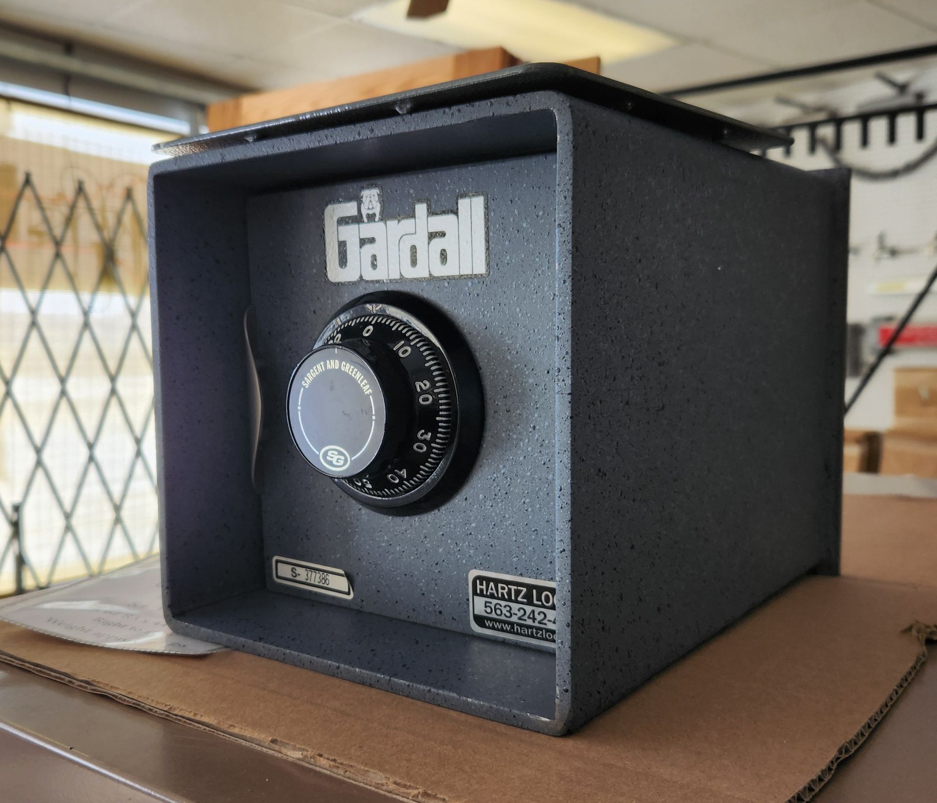 A dark gray Gardall floor safe with a mechanical combination dial sitting on a cardboard surface in an indoor setting.