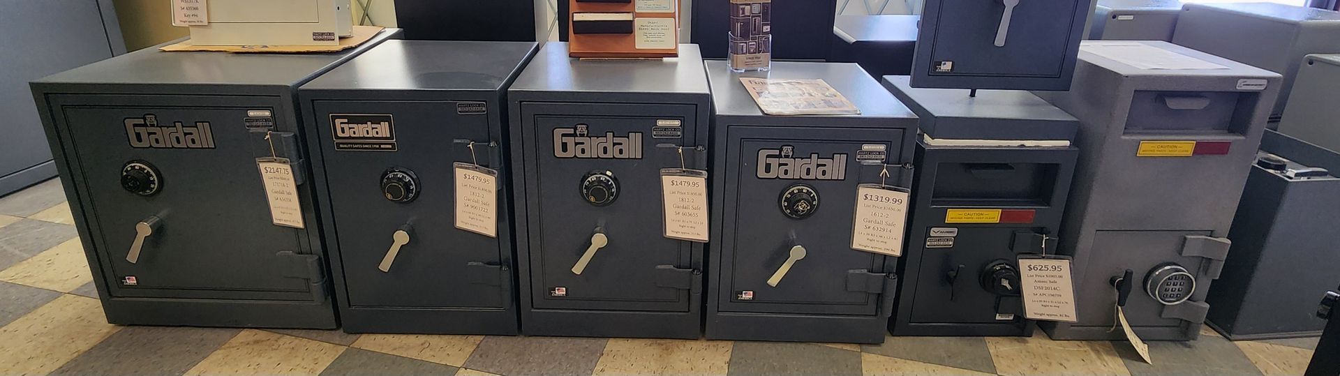 A row of six gray commercial safes of varying styles and sizes arranged side-by-side on a tiled floor.