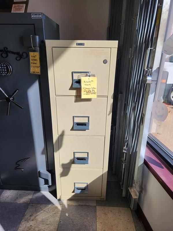 Tan four-drawer filing cabinet next to a metal gate and a dark gray safe. A sign is on the cabinet.