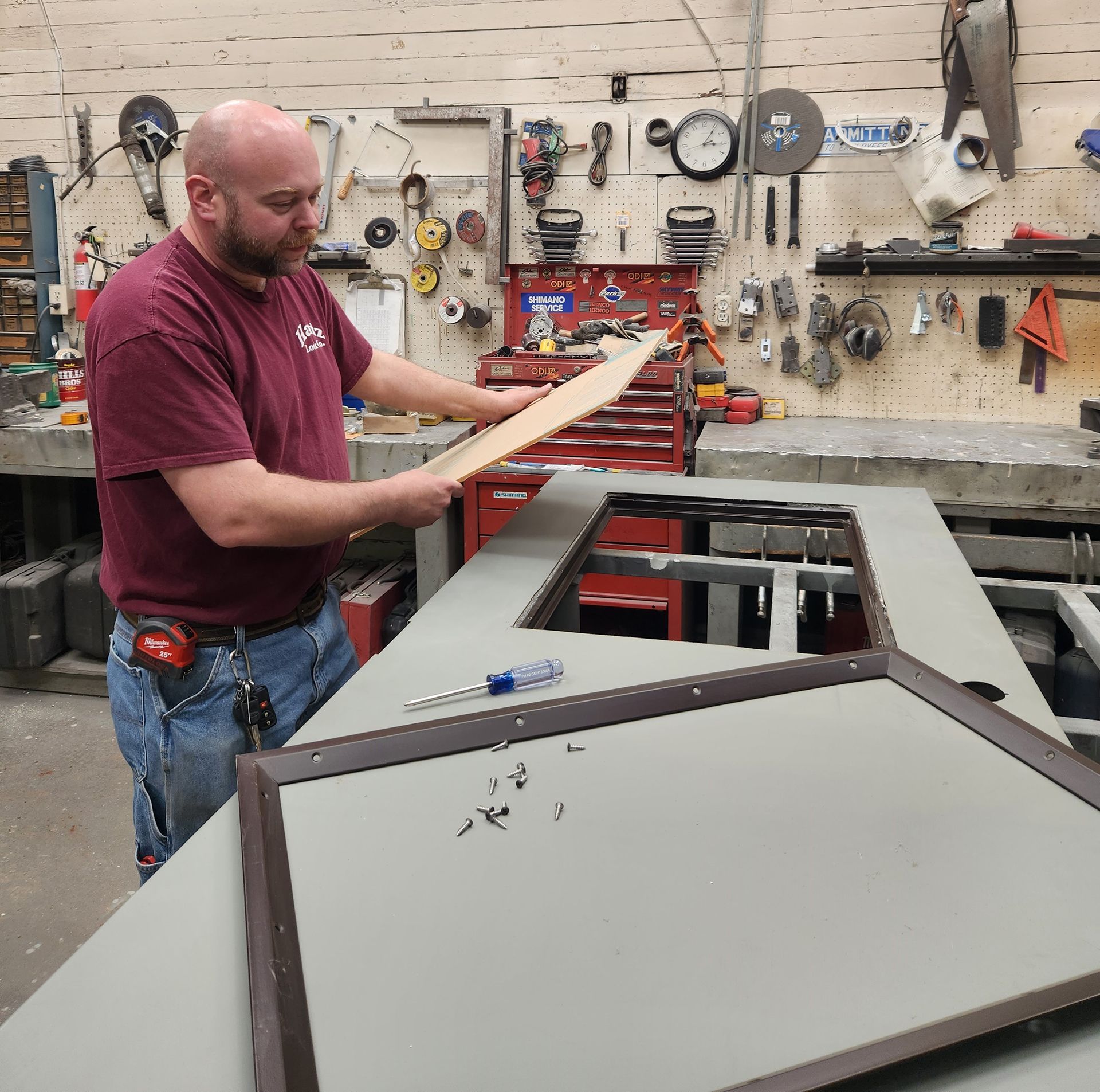 Man in a shop fitting wood into a frame, surrounded by tools and equipment.