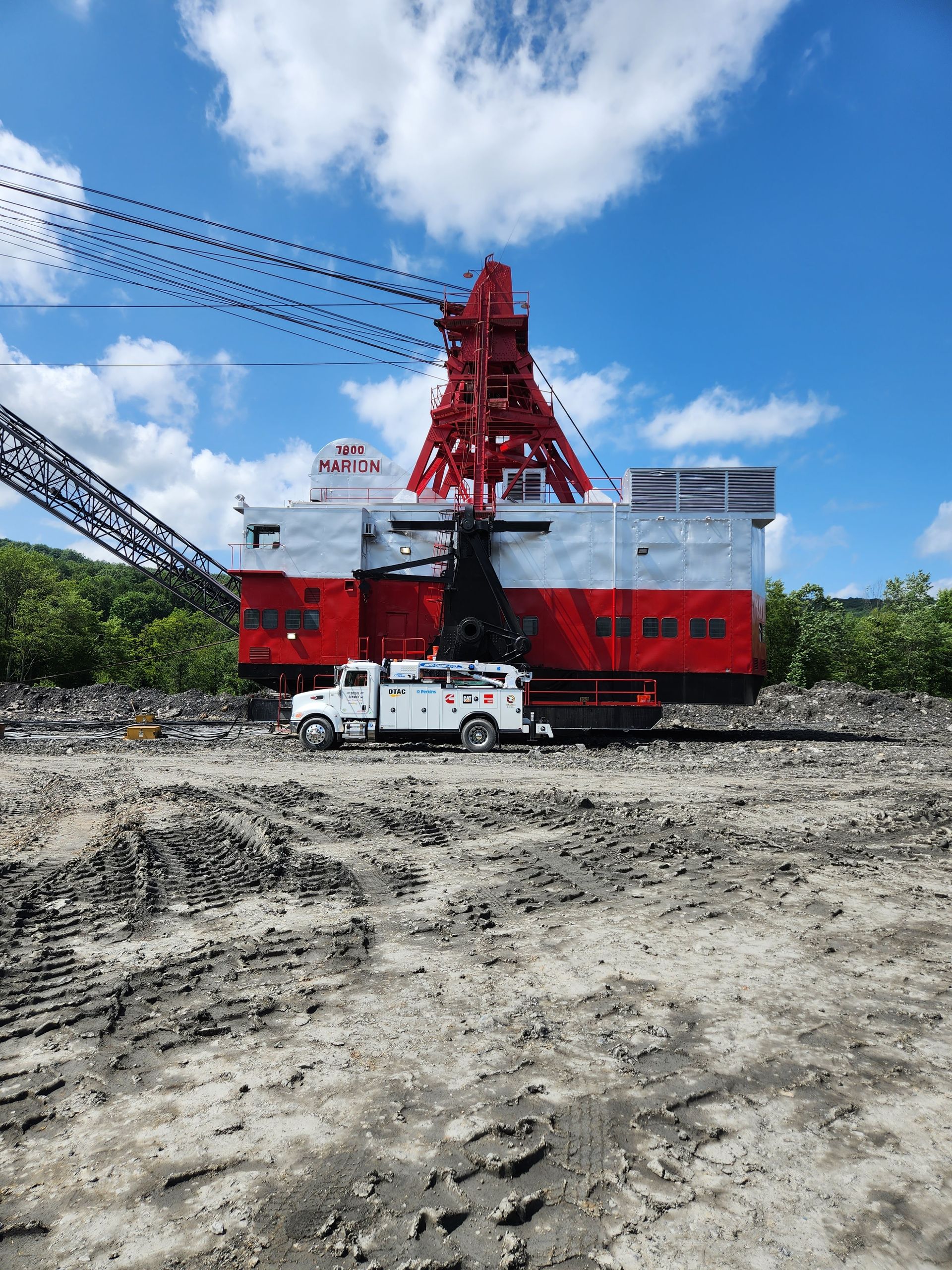 Large red and white mining excavator with support truck on gray earth under blue sky.