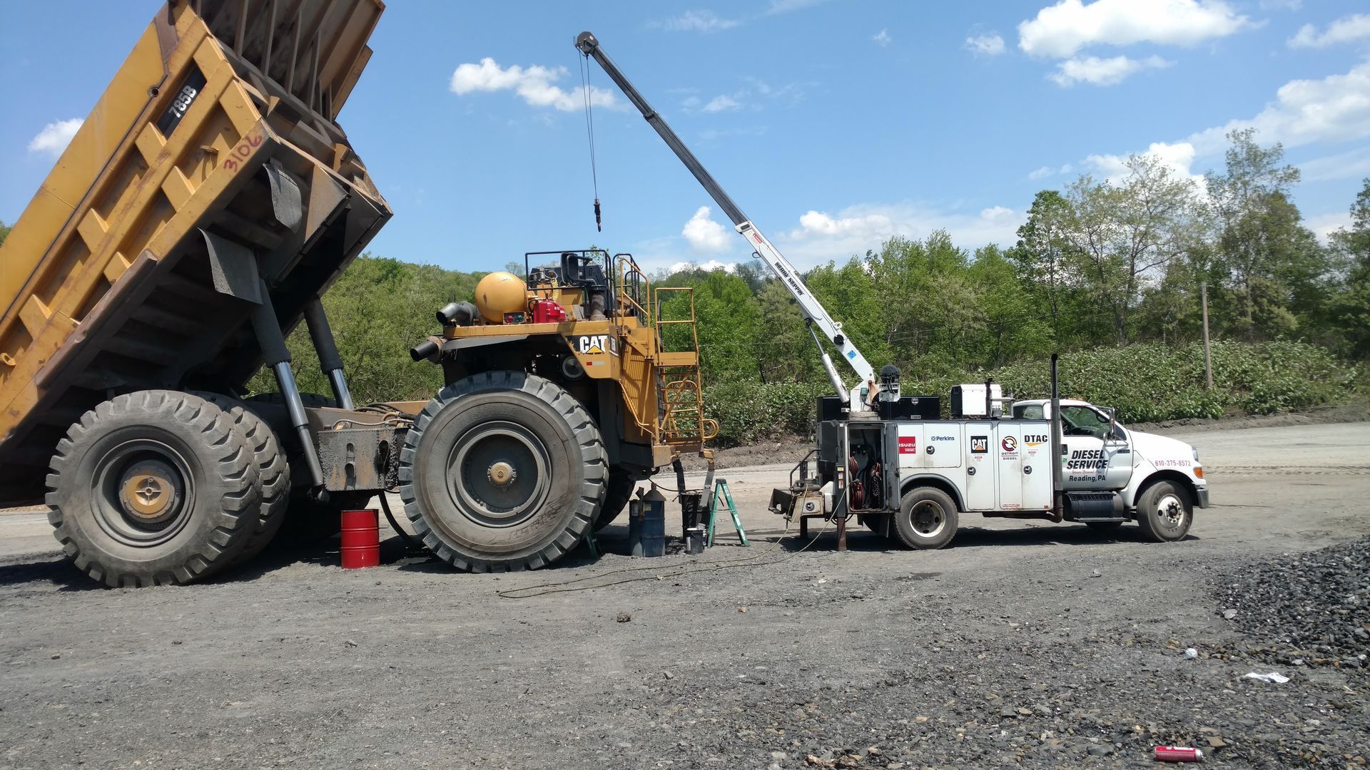 A large dump truck being serviced by a white service truck with a crane in a gravel area.