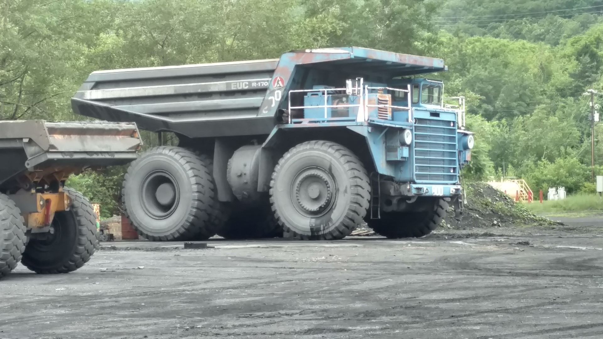 Large blue mining dump truck on a surface parking lot. Tires are black, setting is outdoors, next to a smaller, similar truck.
