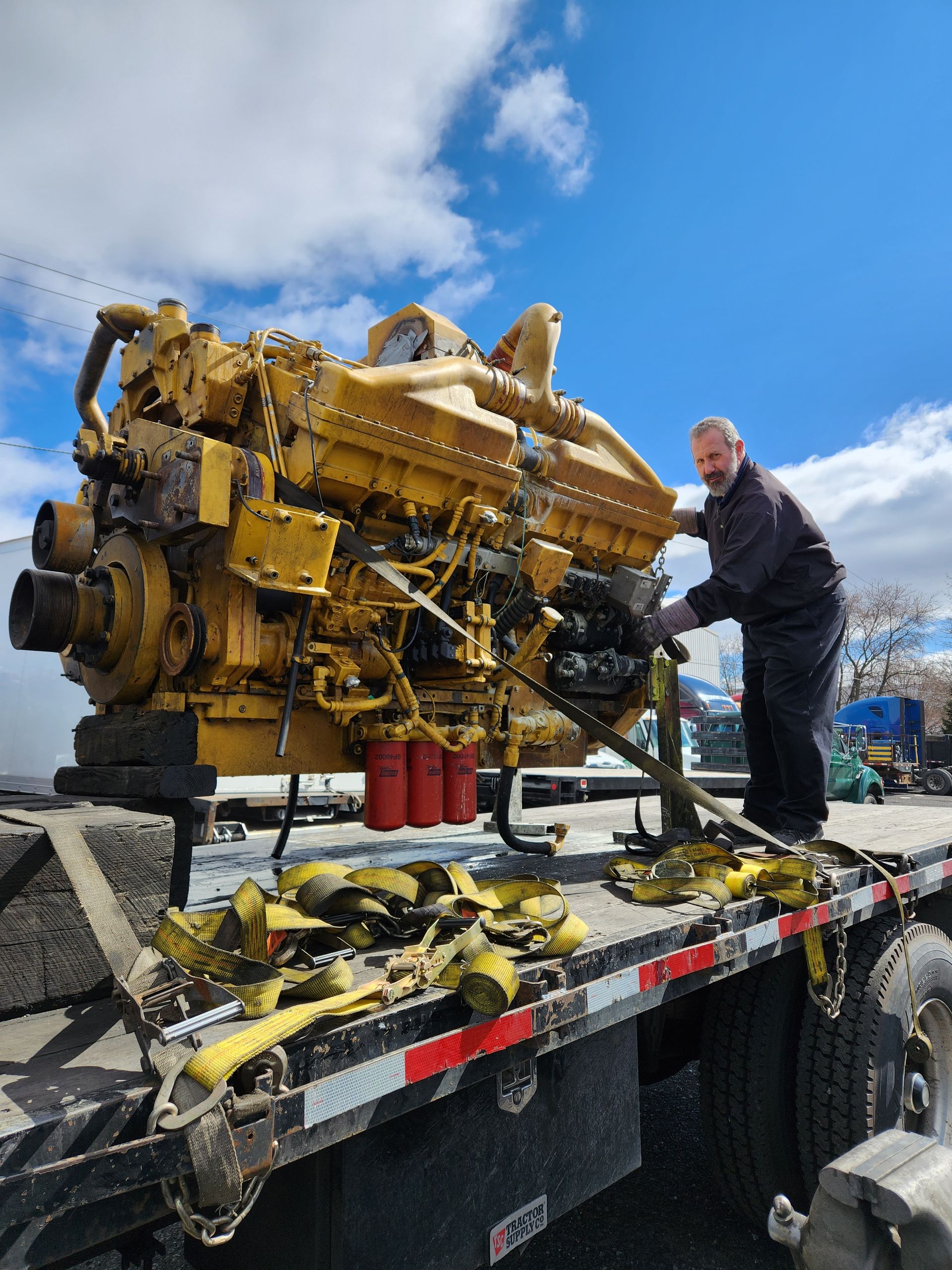 A large yellow engine secured to a flatbed truck by straps. A man stands nearby. Blue sky.