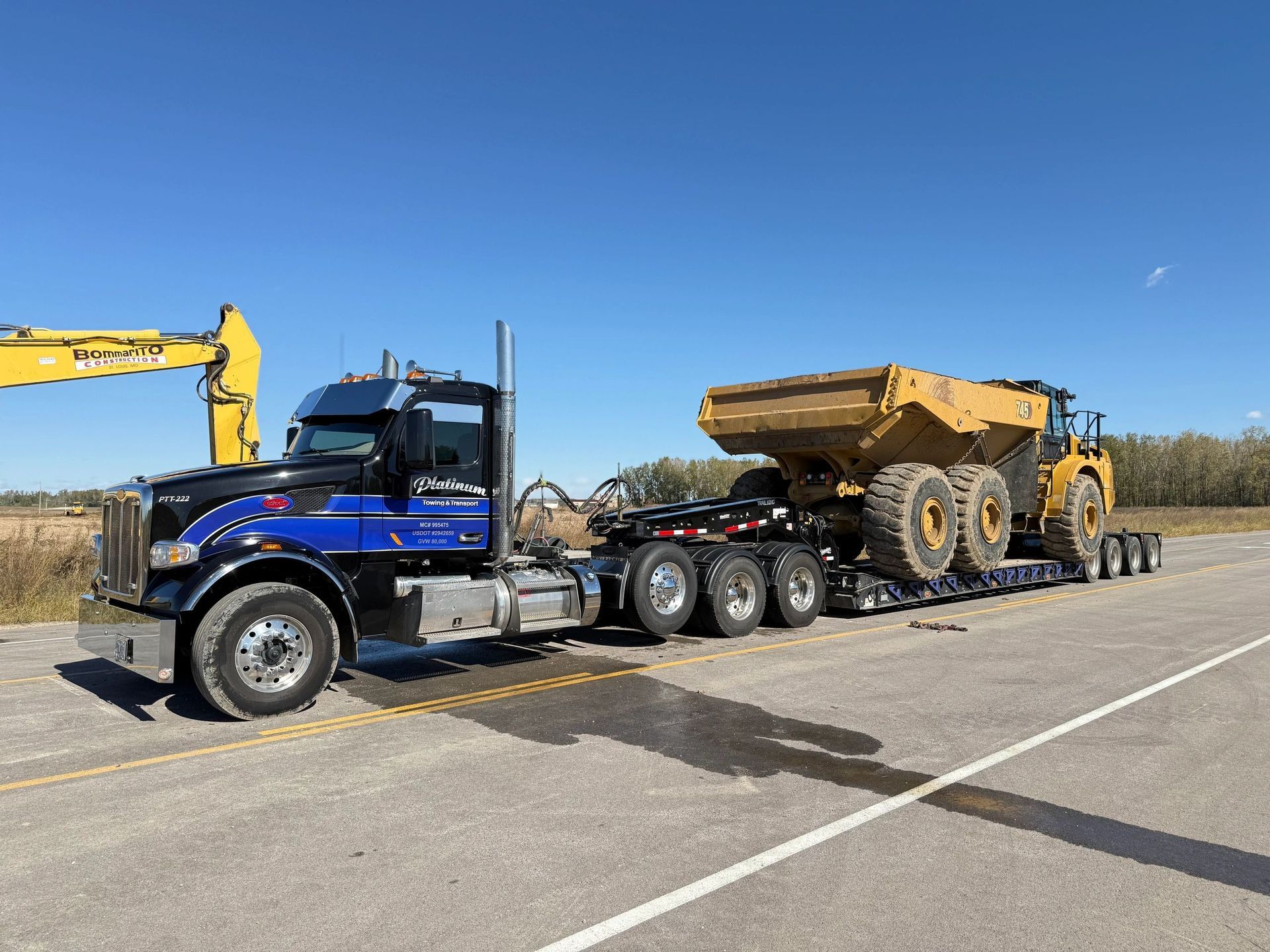 Blue and black semi-truck hauling a yellow dump truck on a lowboy trailer, parked on pavement, clear sky.