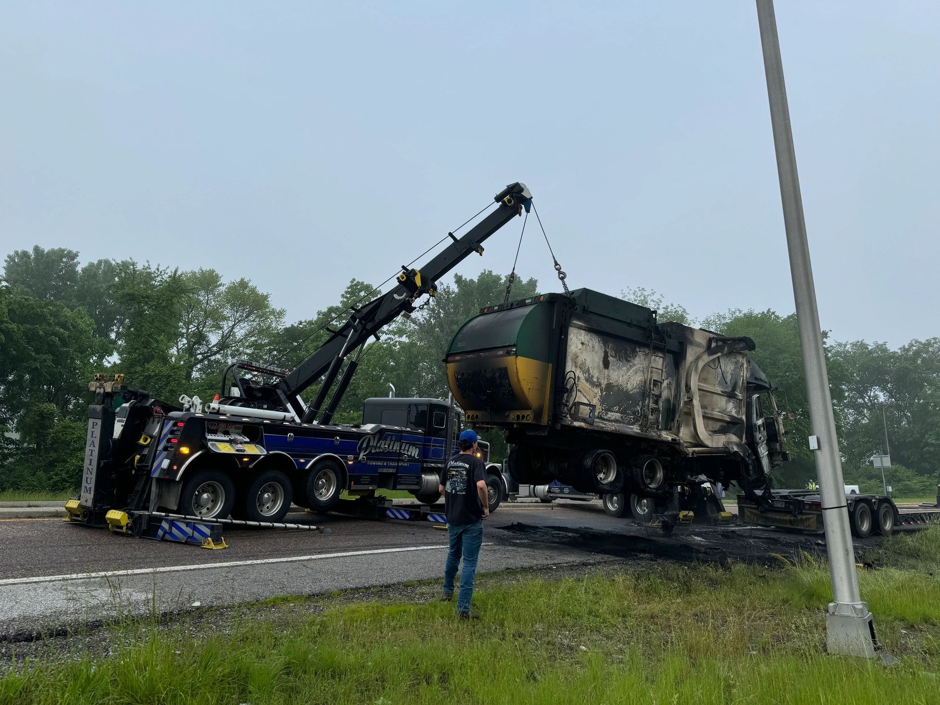 A tow truck lifts a heavily damaged garbage truck from a highway. The truck's body is charred. A man watches.
