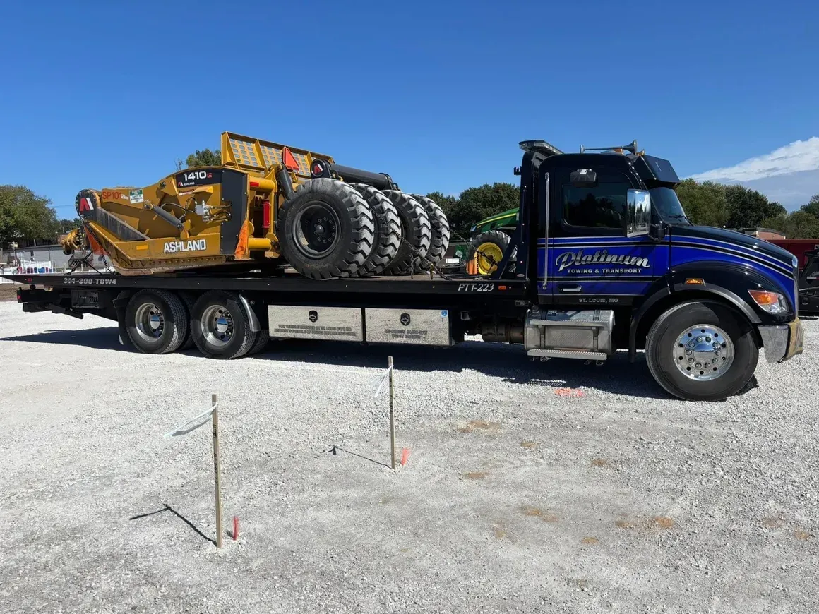 Flatbed tow truck carrying a large yellow and black industrial machine on a gravel lot under a blue sky.