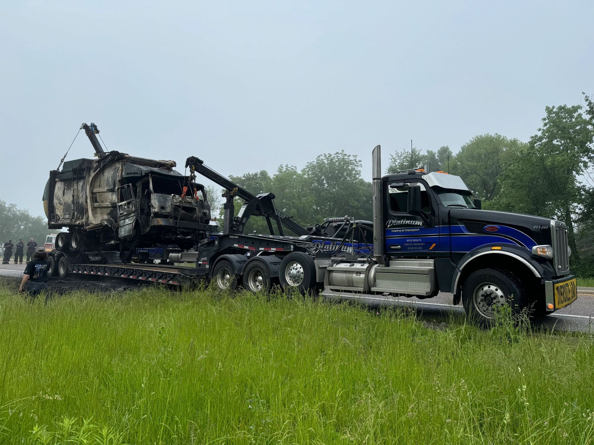 A tow truck hauling a heavily damaged vehicle on a flatbed trailer next to a grassy roadside.