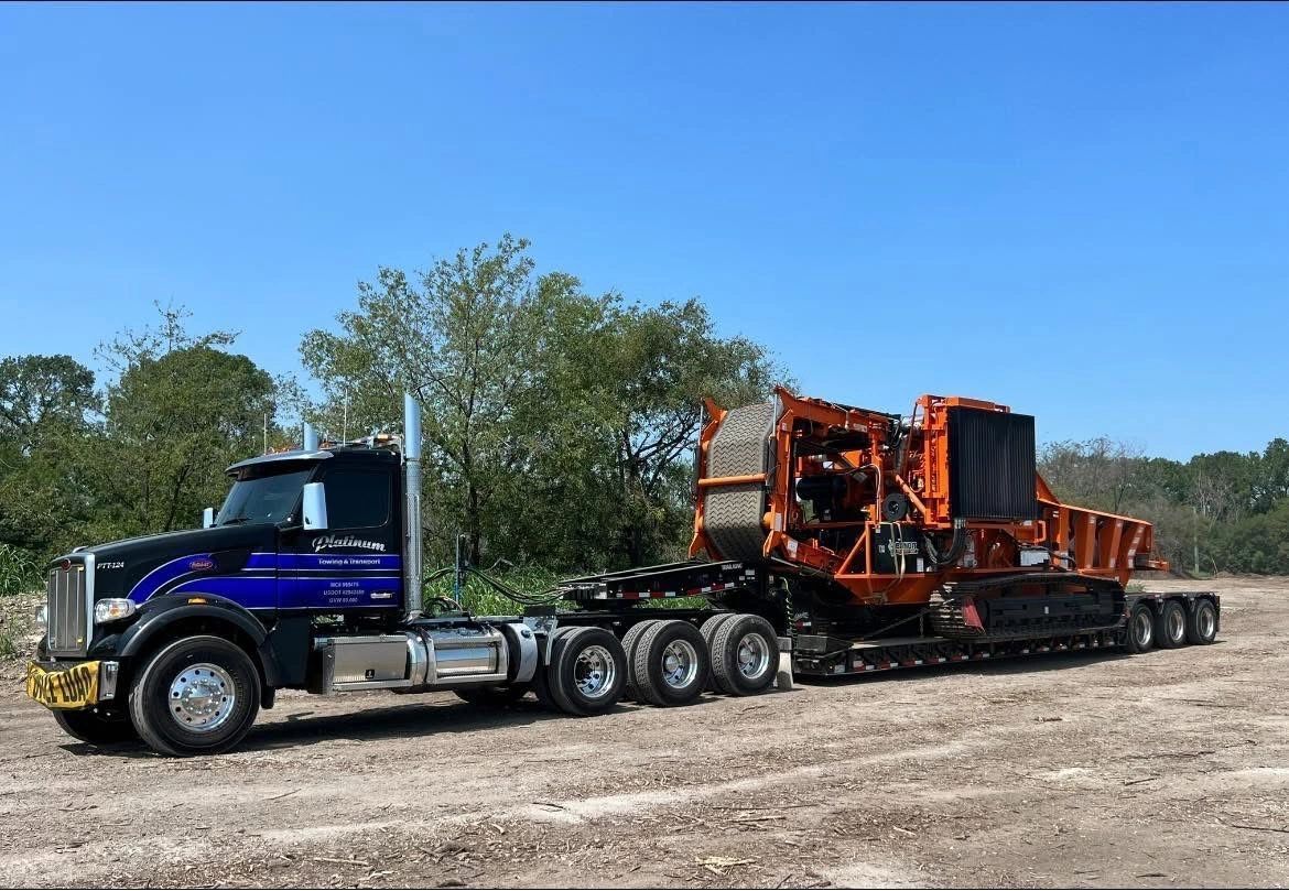 Black semi-truck hauling an orange industrial machine on a flatbed trailer on a dirt road under a blue sky.