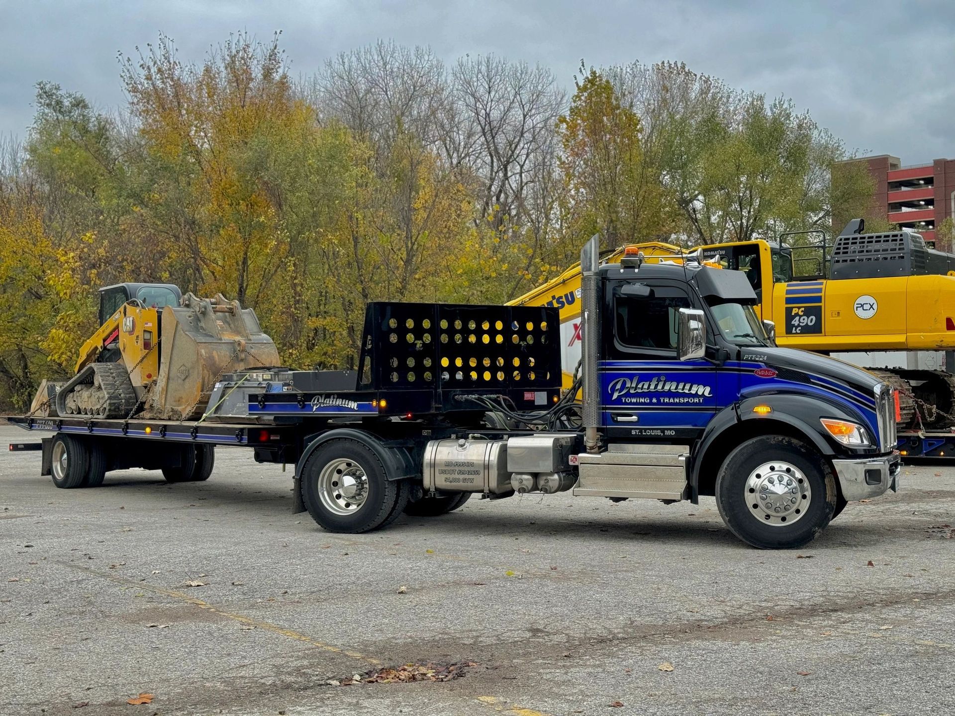 Blue truck with flatbed trailer carrying a small excavator and a large rock, parked outdoors.