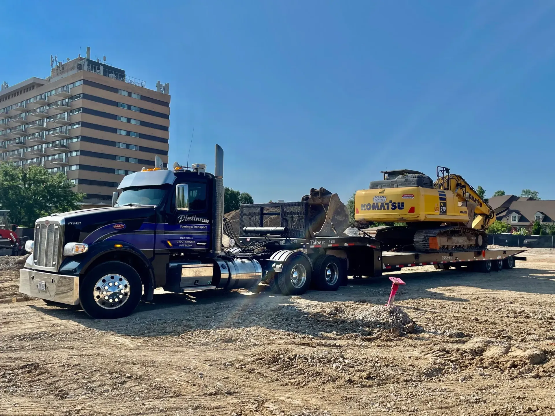 Dark blue semi-truck with a yellow excavator on a trailer at a construction site on a sunny day.