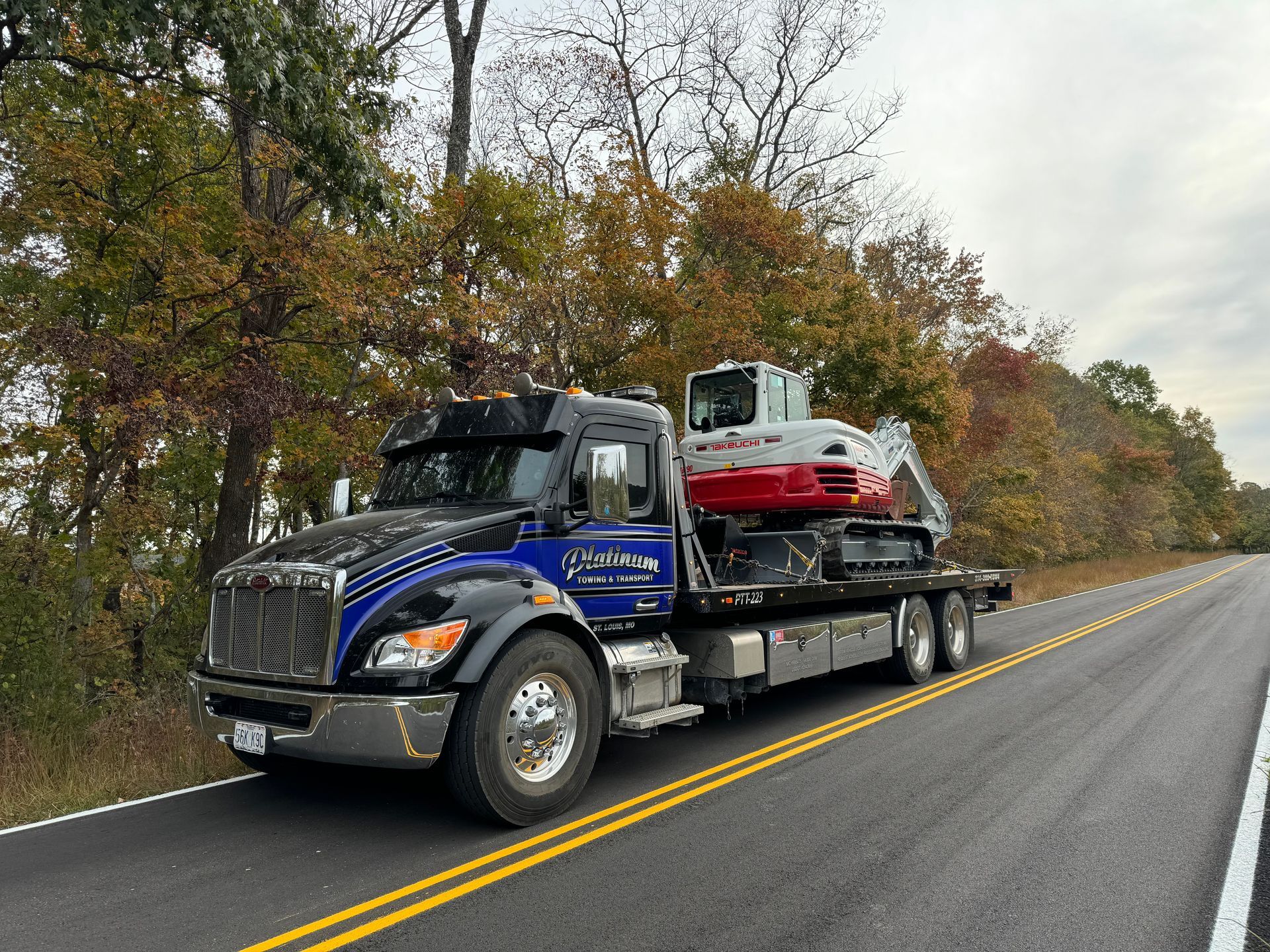 A flatbed truck transporting a small excavator on a road lined with trees.