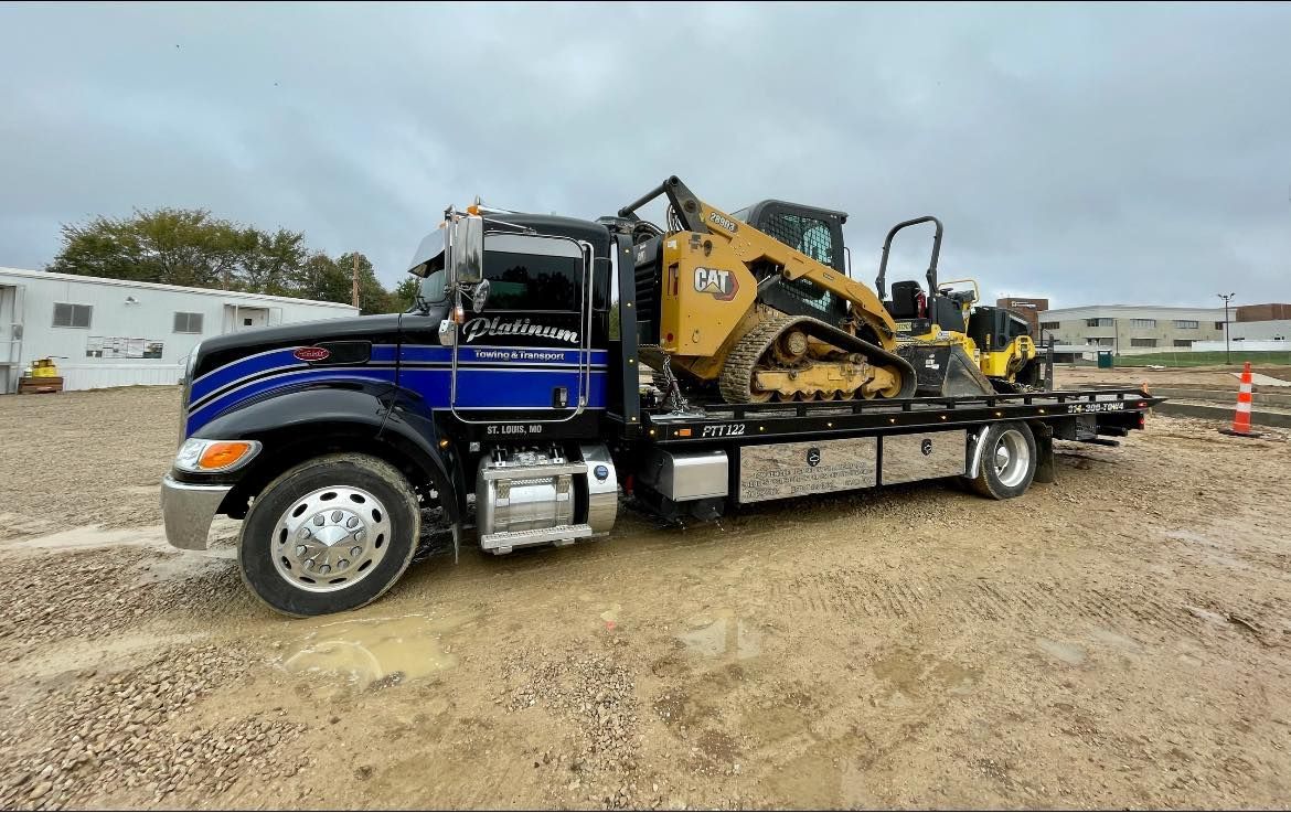 A flatbed tow truck with a yellow Caterpillar track loader on the bed, parked on gravel.
