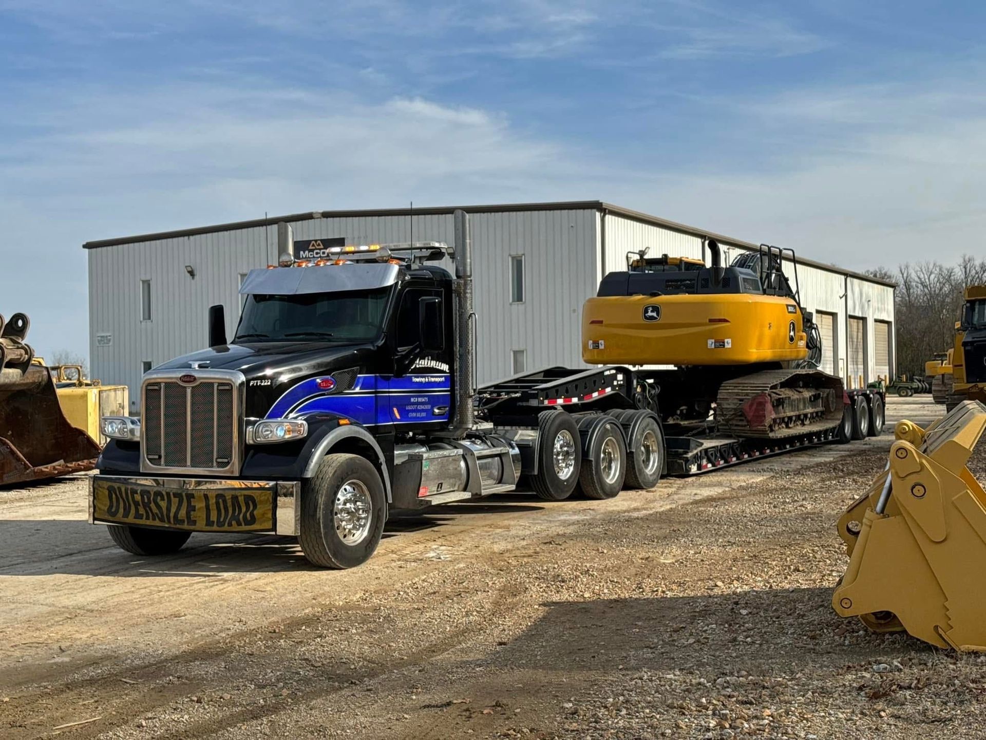 A semi-truck hauling a yellow excavator on a flatbed trailer, parked in front of a white building.