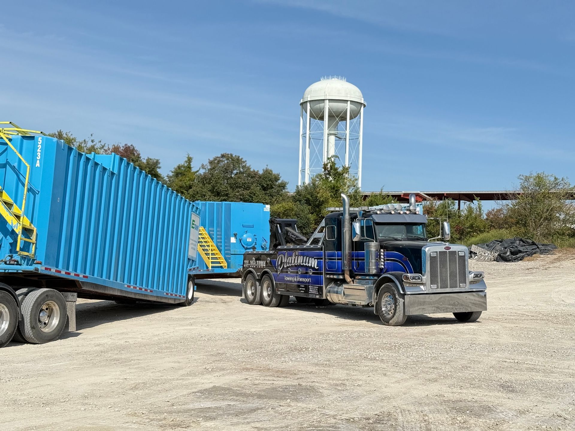 Blue semi-truck towing a large, blue container. Water tower in background under a blue sky.