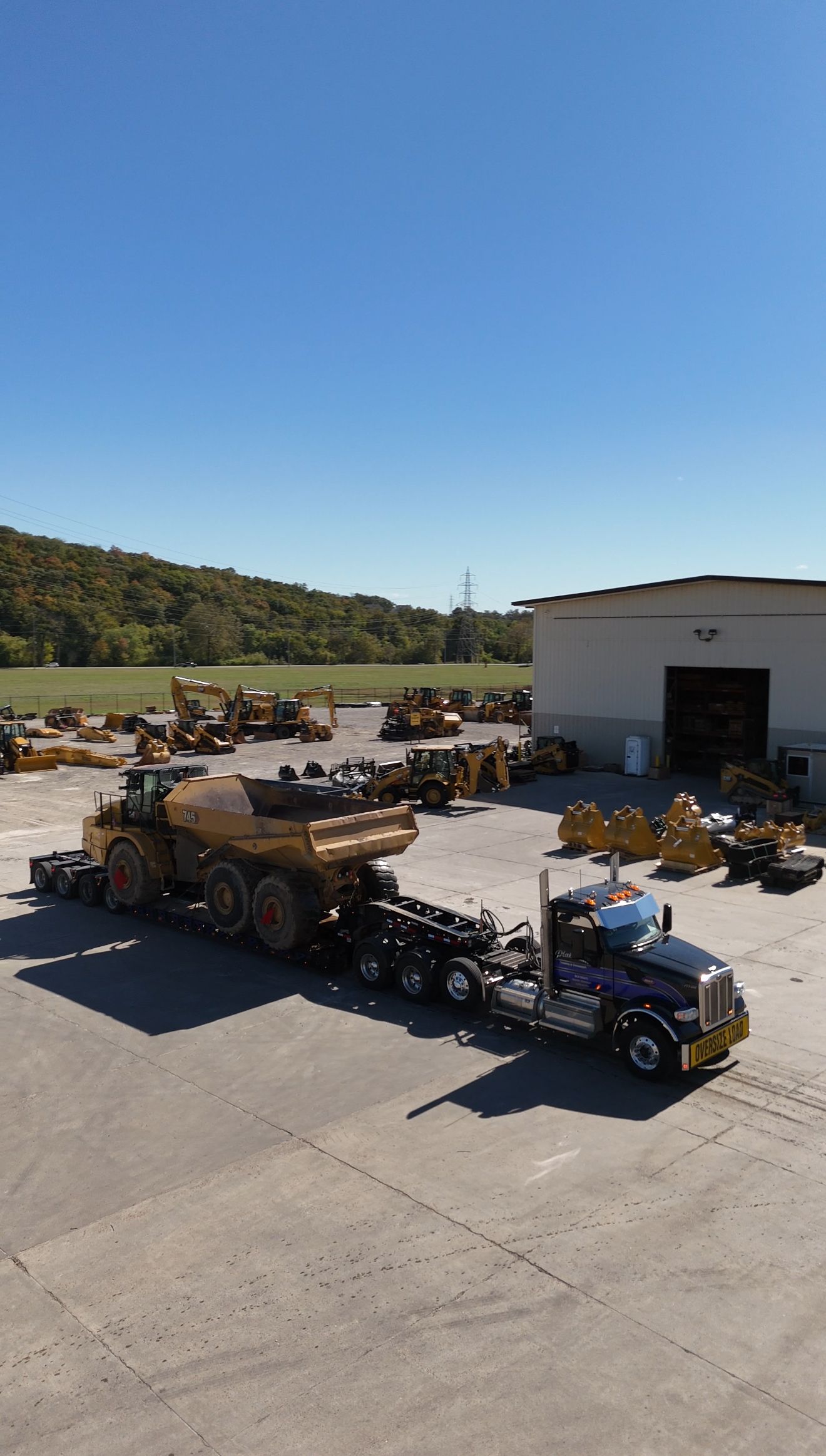 Semi-truck hauling heavy machinery on a trailer in a lot, sunny day.