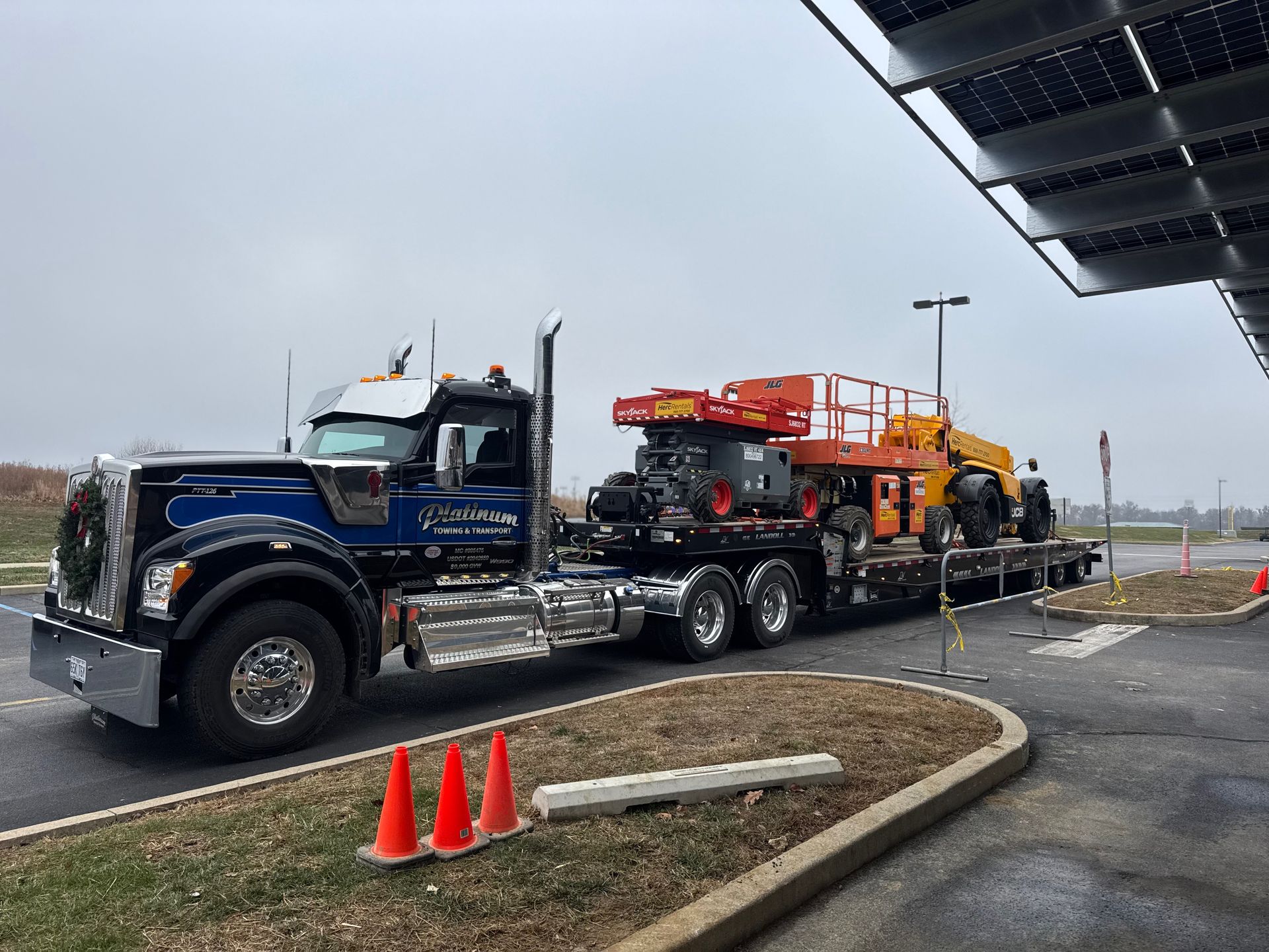 Semi-truck hauling construction equipment under a solar panel canopy, next to a curb.