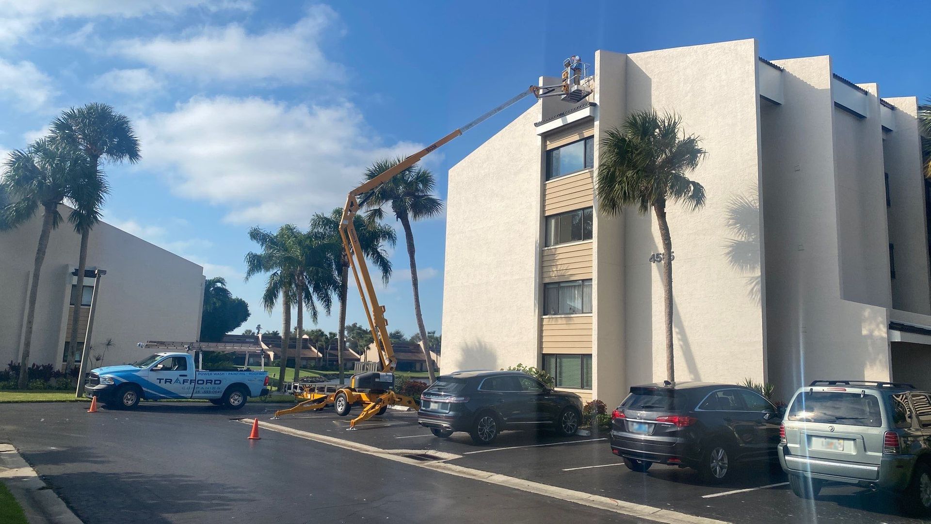 Construction work on a multistory building. A crane extends to the roof, vehicles and palm trees surround it.