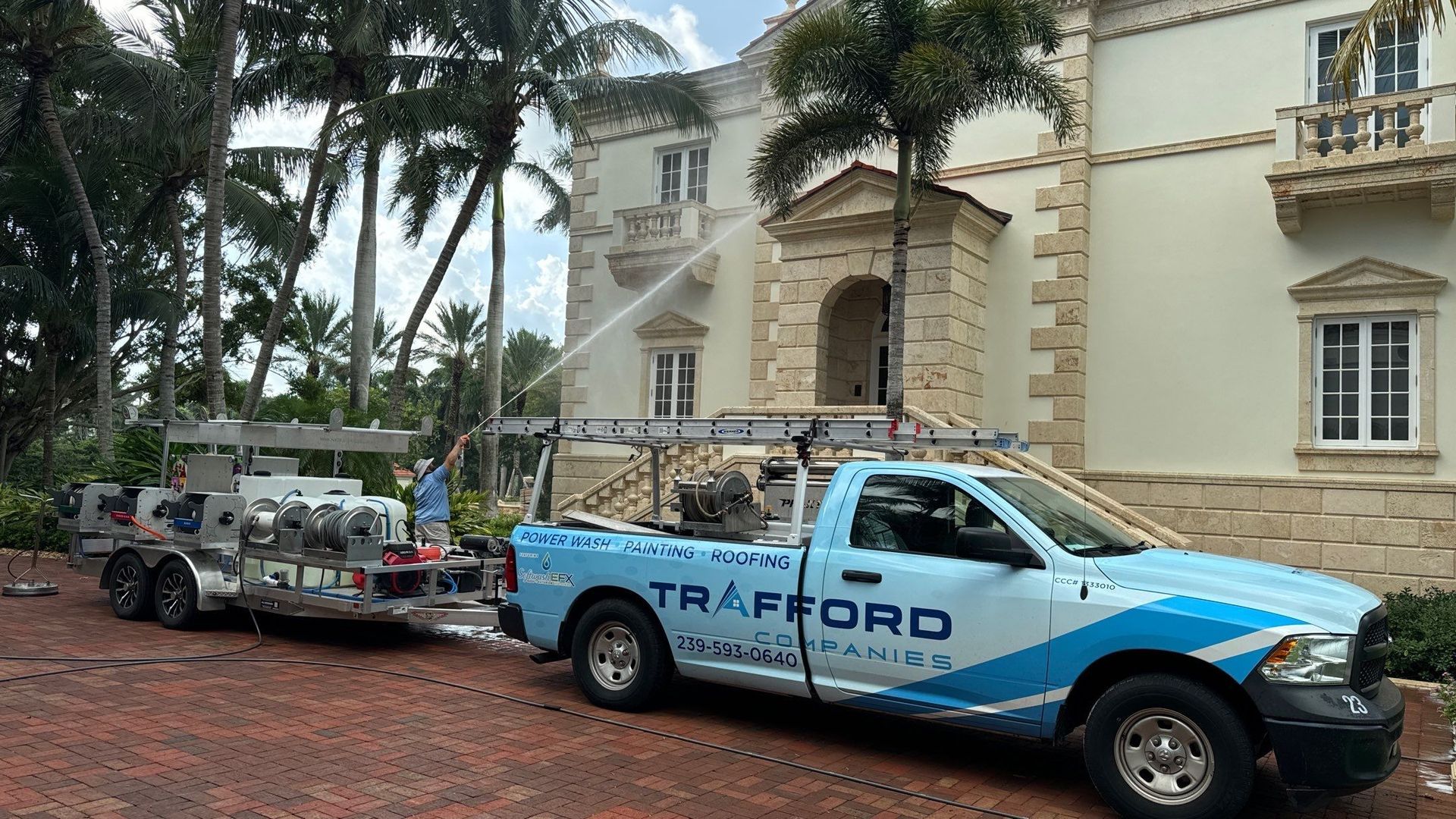 A blue truck with TRAFFORD logo washes a large cream-colored building. Trailer attached. Palm trees surround.
