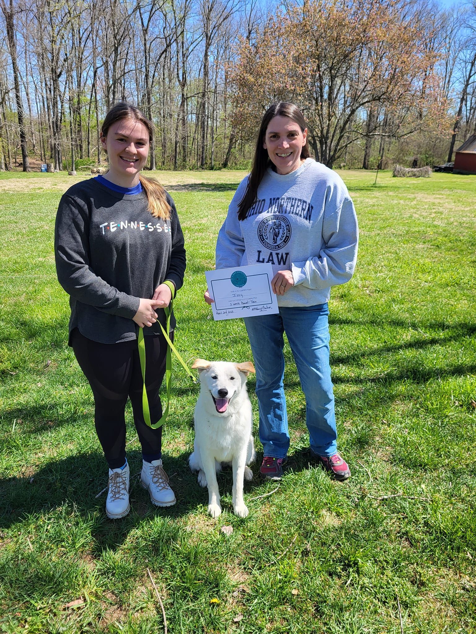 Two women are standing next to a white dog in a grassy field