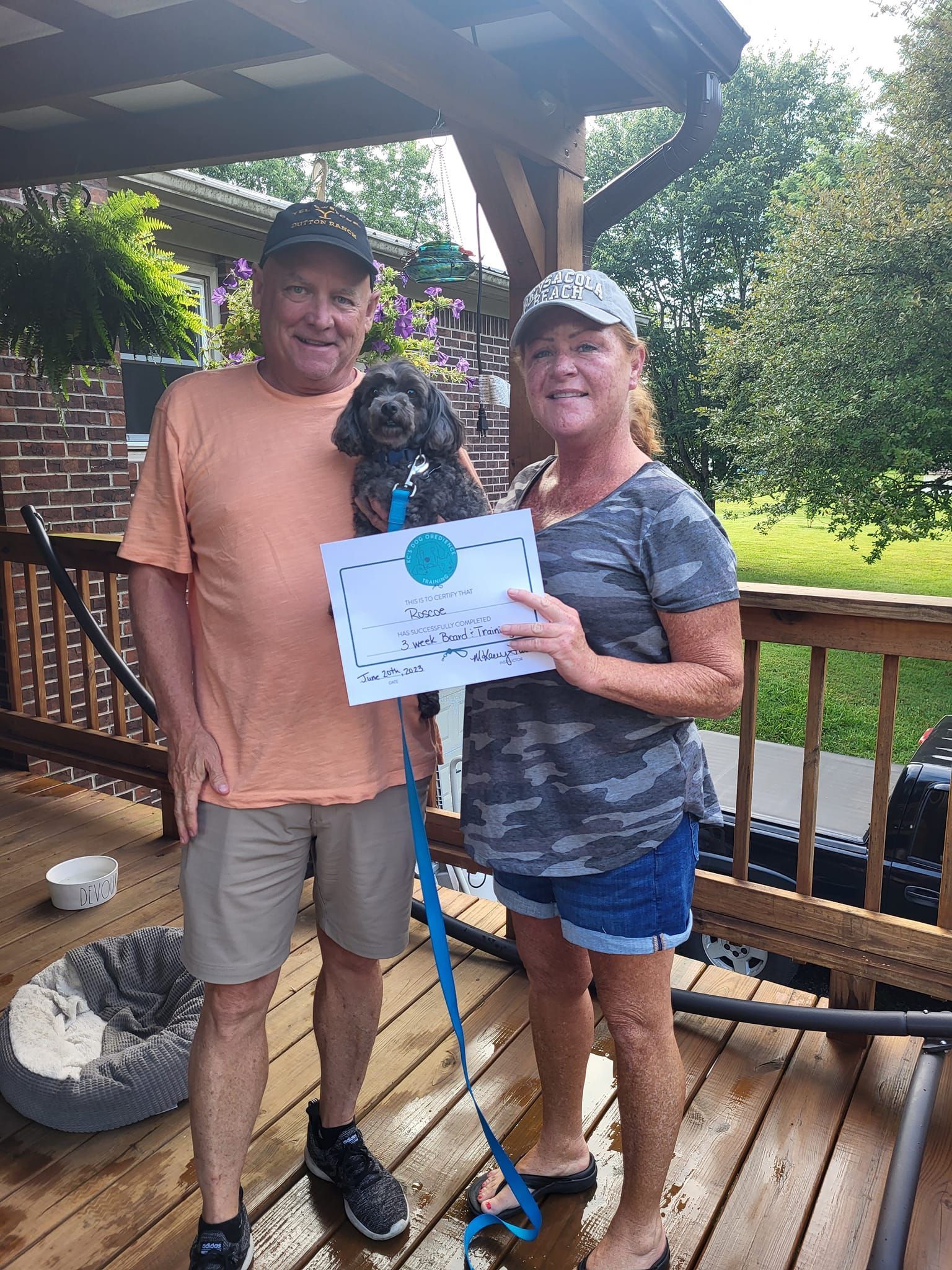 A man and a woman are standing on a deck with a dog and holding a certificate