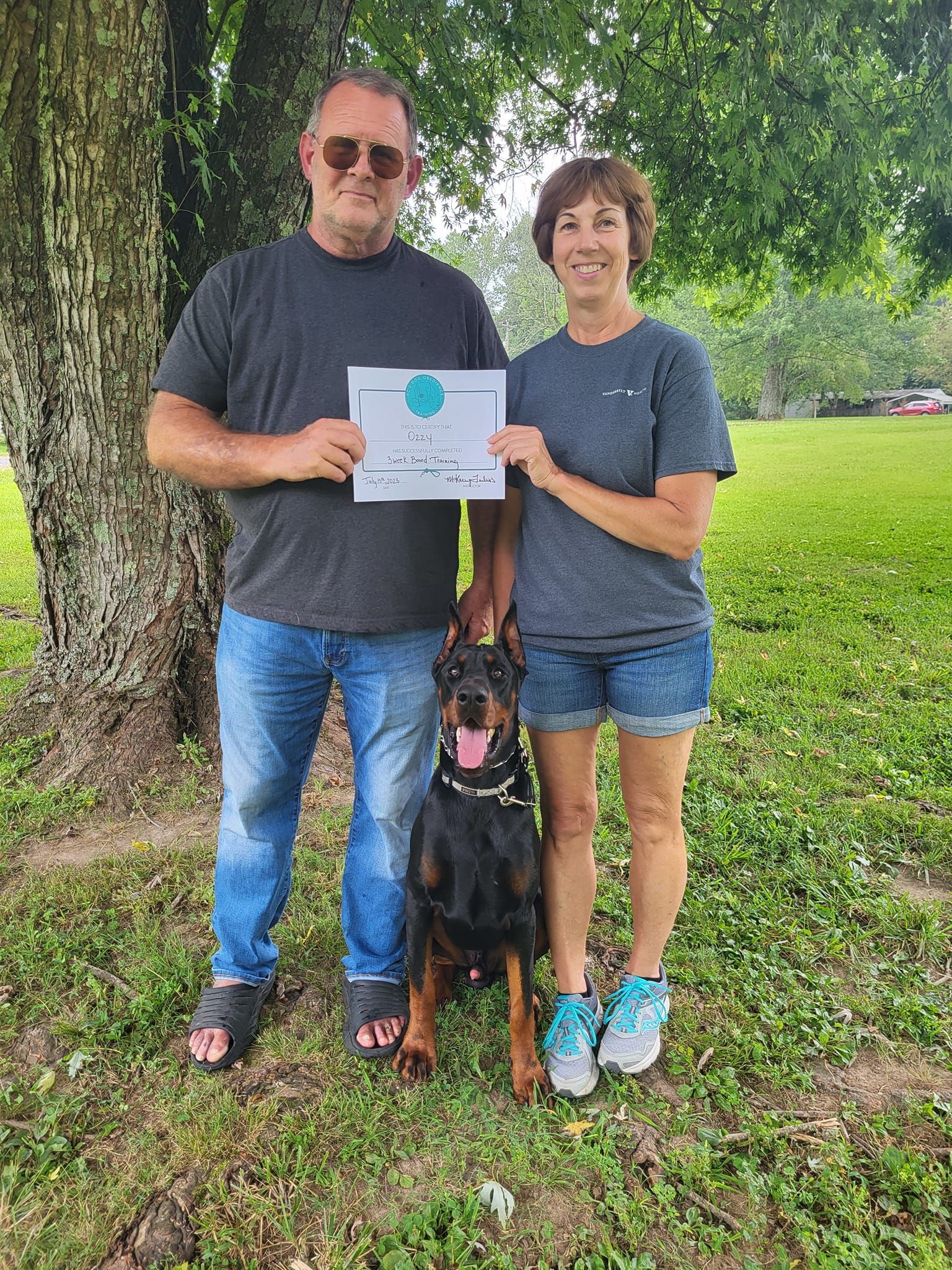 A man and woman are standing next to a dog in a park holding a certificate