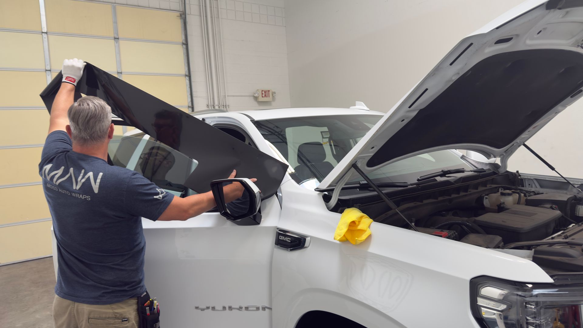 Man removing a windshield film from a white pickup truck in a garage, hood open.