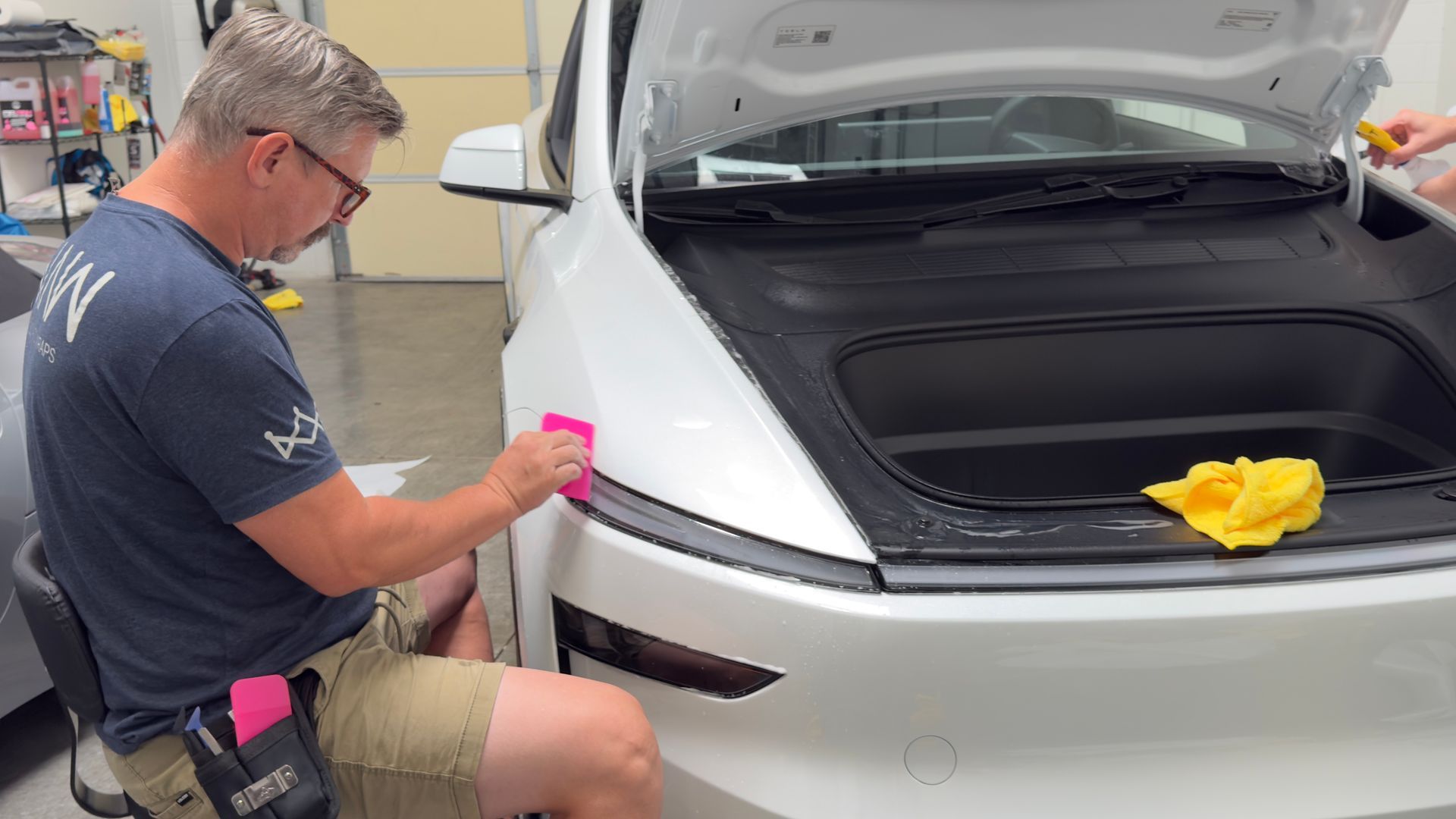 Technician polishing the front bumper of a white car with the hood open in a workshop